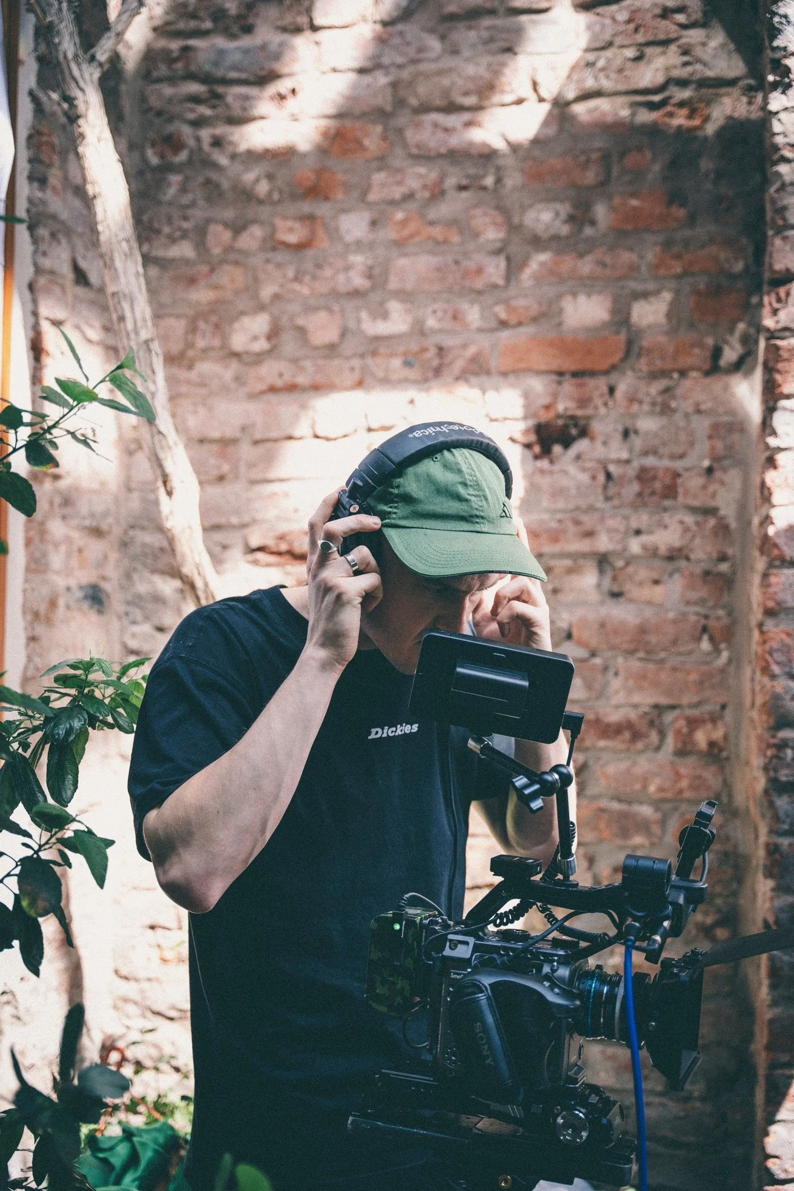 Person wearing a green cap and black shirt, adjusting a professional camera setup in an indoor space with a brick wall background and some green plants.