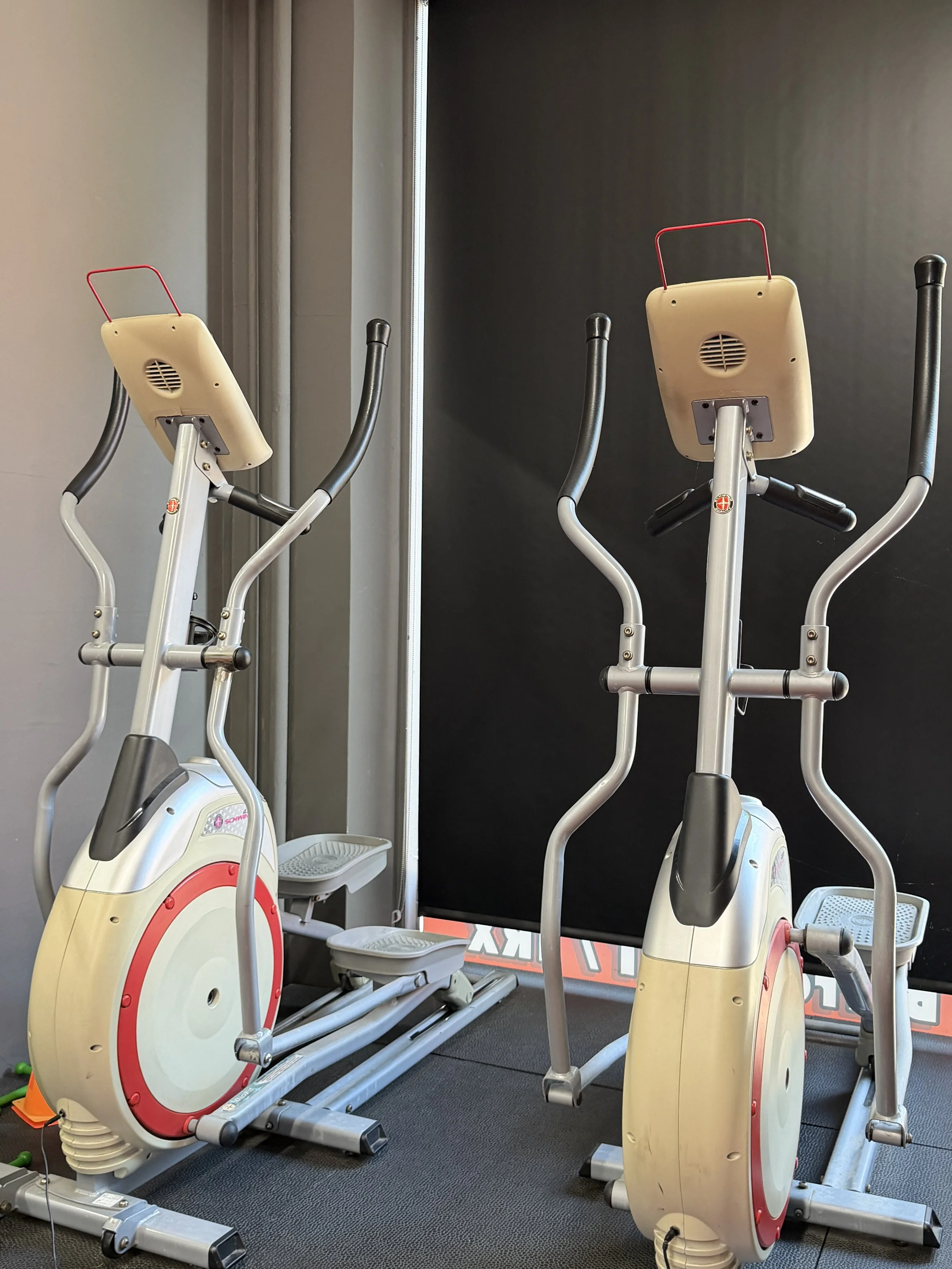 Two yellow elliptical exercise bikes with black handles and red and white flywheels, positioned next to each other in a gym or fitness studio with black and gray walls.