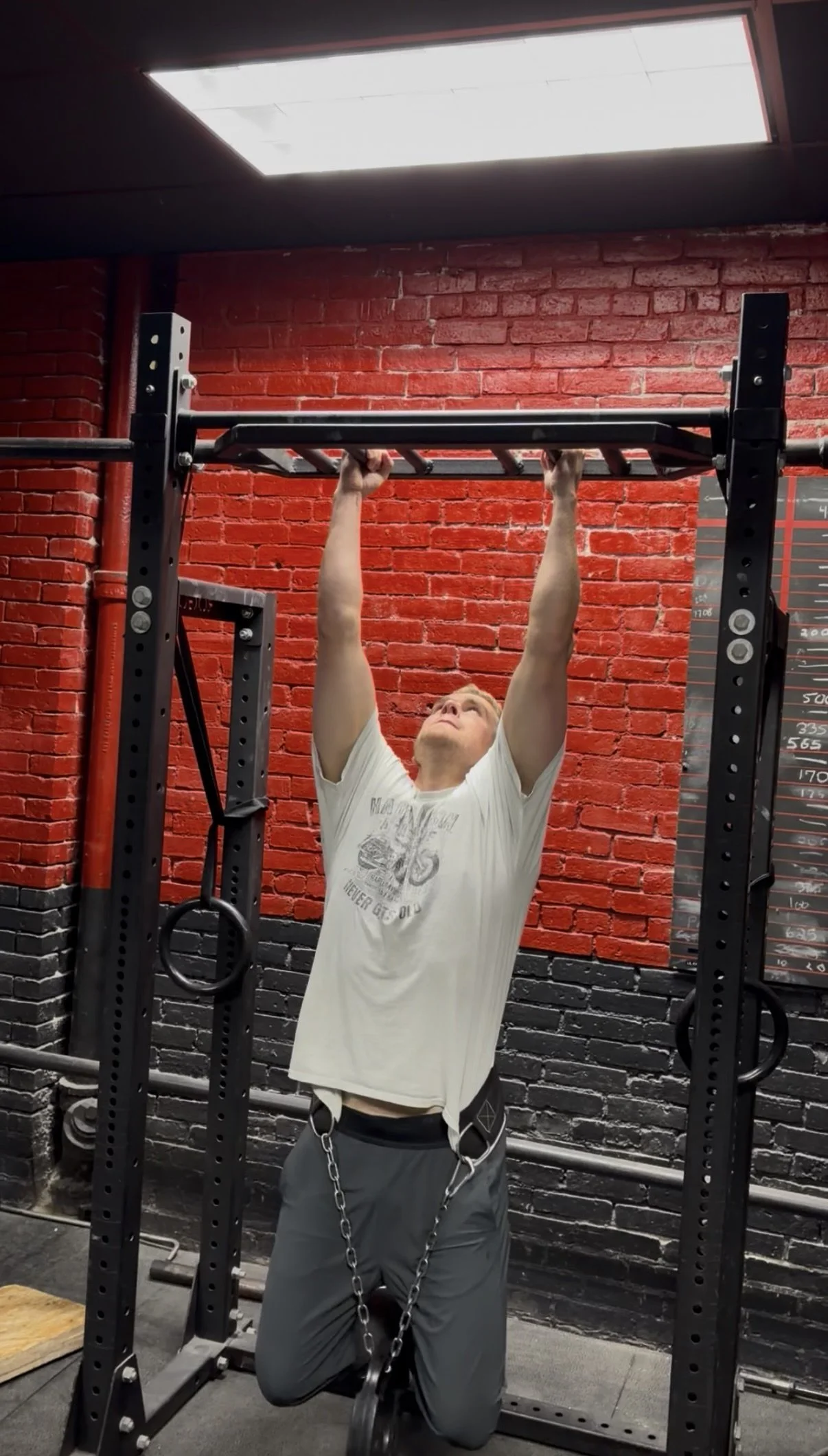 A man with a chain around his legs and a white t-shirt is hanging from a pull-up bar in a gym with red brick walls.