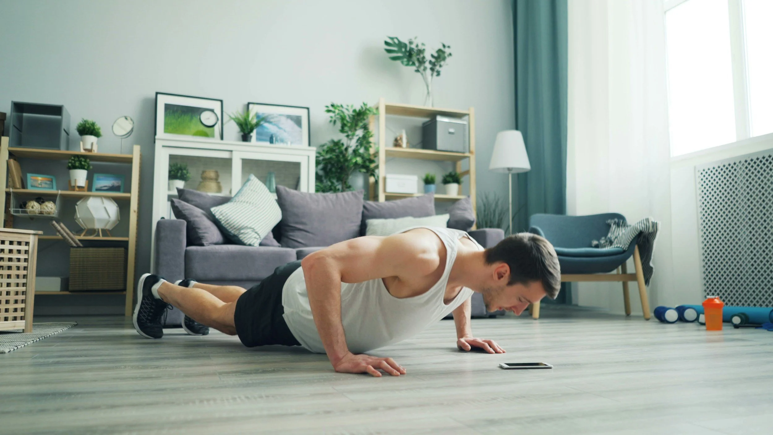 Young man in workout clothes doing push-ups on the floor of a living room with indoor plants, framed pictures, and a gray sofa in the background.