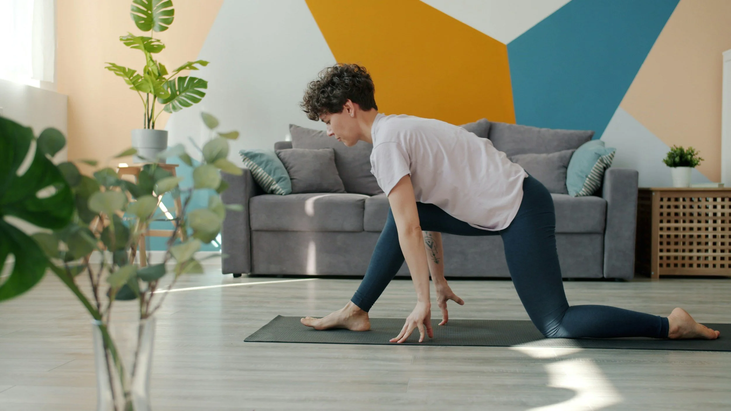 A woman practicing yoga indoors, kneeling on a yoga mat in front of a gray couch and colorful geometric wall. There are plants on the side tables and in the foreground.