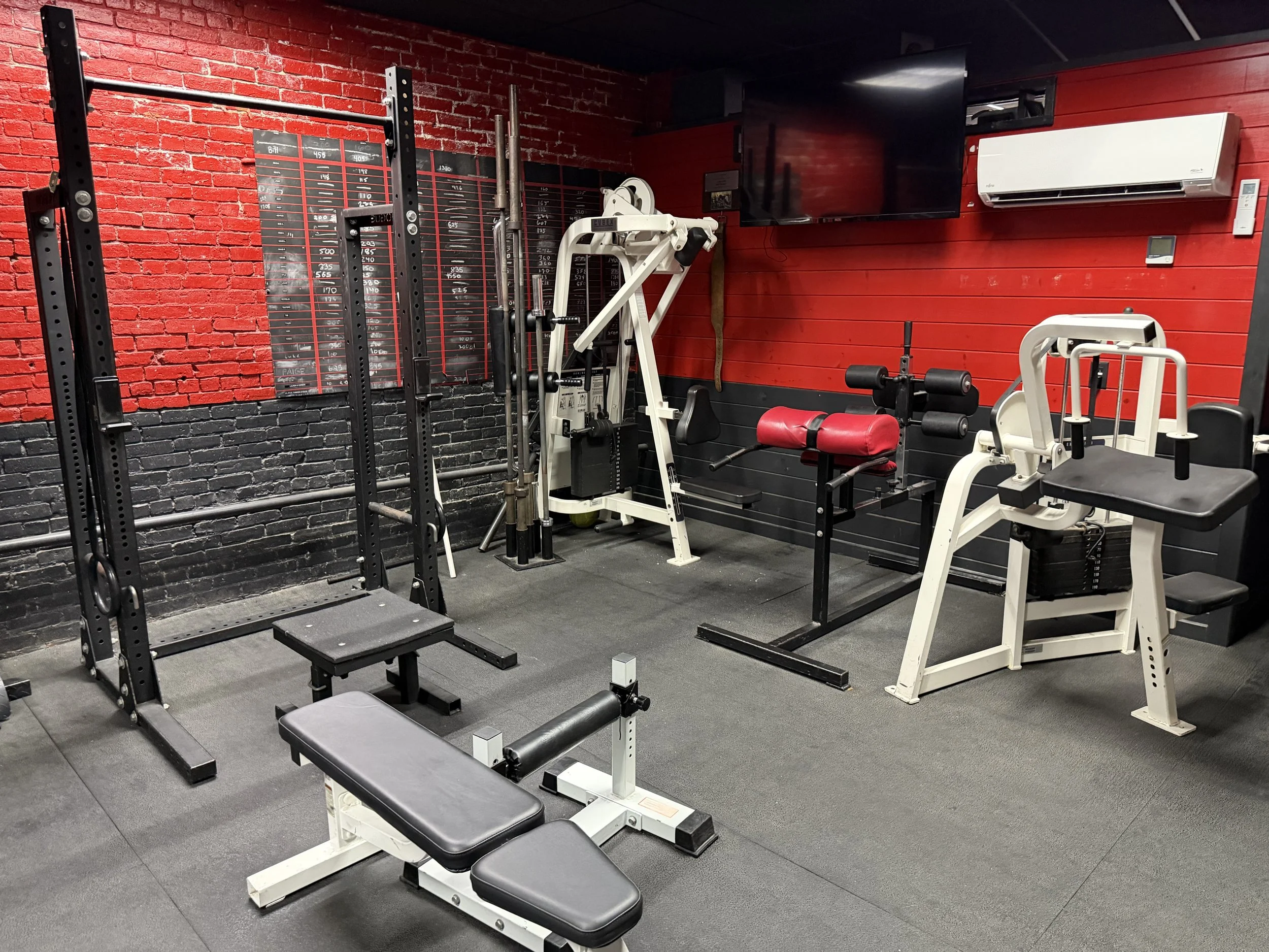 A small gym with black, red, and white workout equipment including a bench press, smith machine, various free weights, and a wall-mounted TV. The walls are painted with a red brick and red panel design, and there is a black carpeted floor.