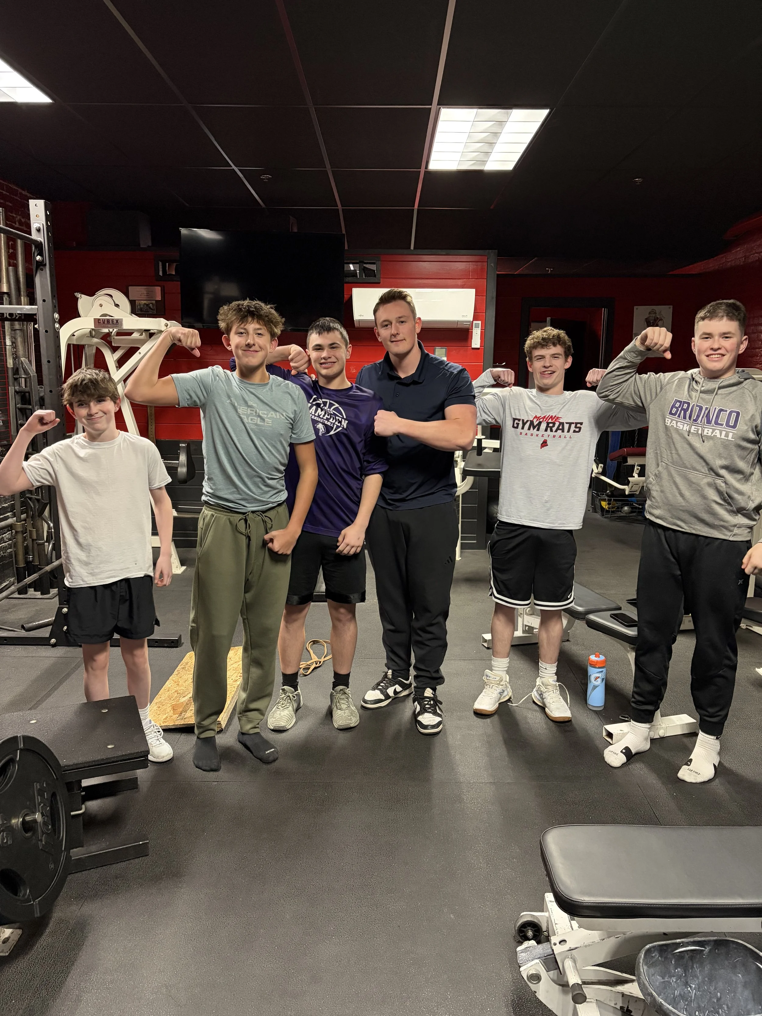 A group of six young men in a gym flexing their muscles and smiling at the camera. They are standing in front of gym equipment and a red wall.