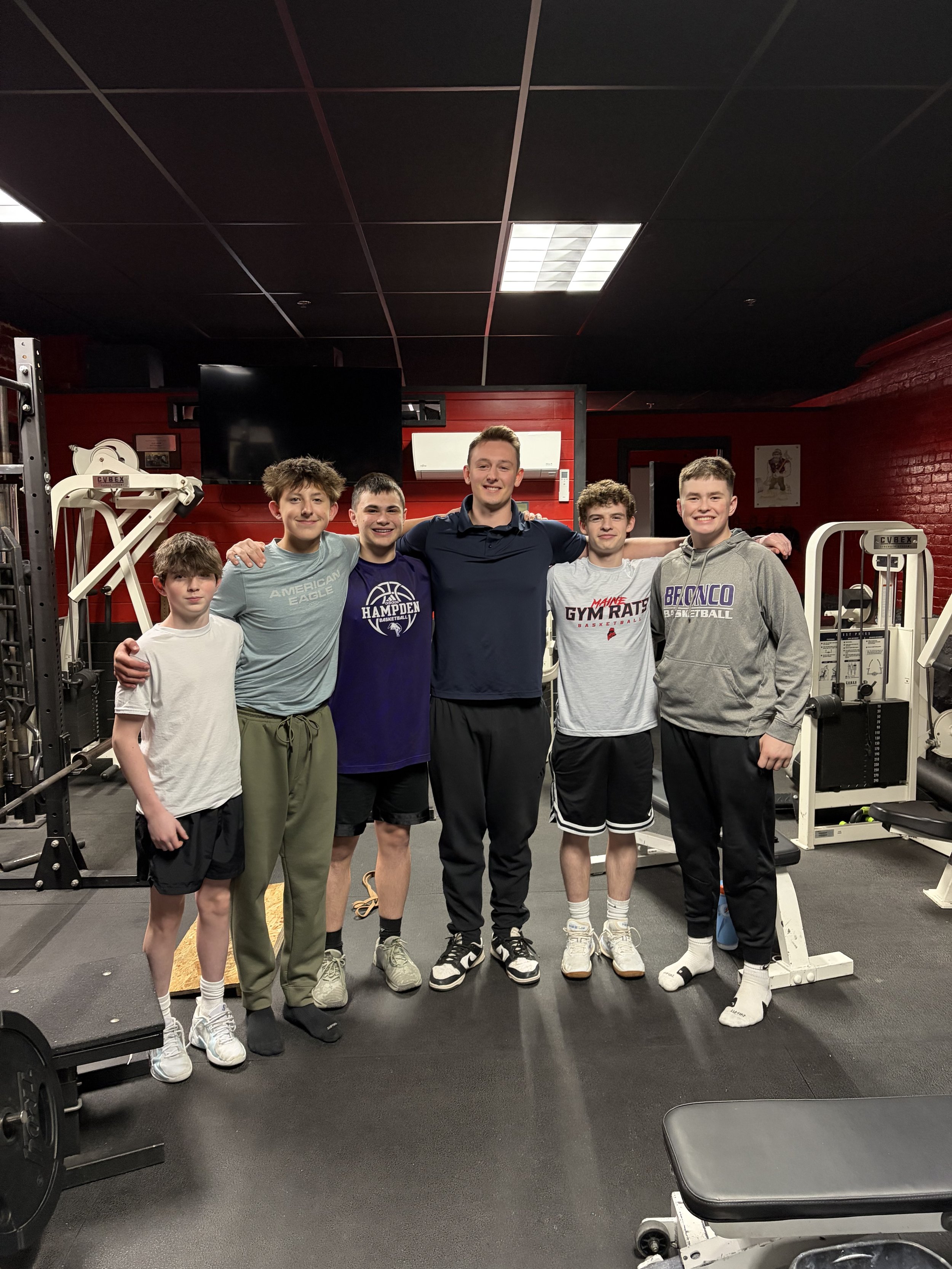 A group of six teenage boys in a gym, standing close together with their arms around each other, smiling at the camera. The gym equipment and black ceiling are visible in the background.