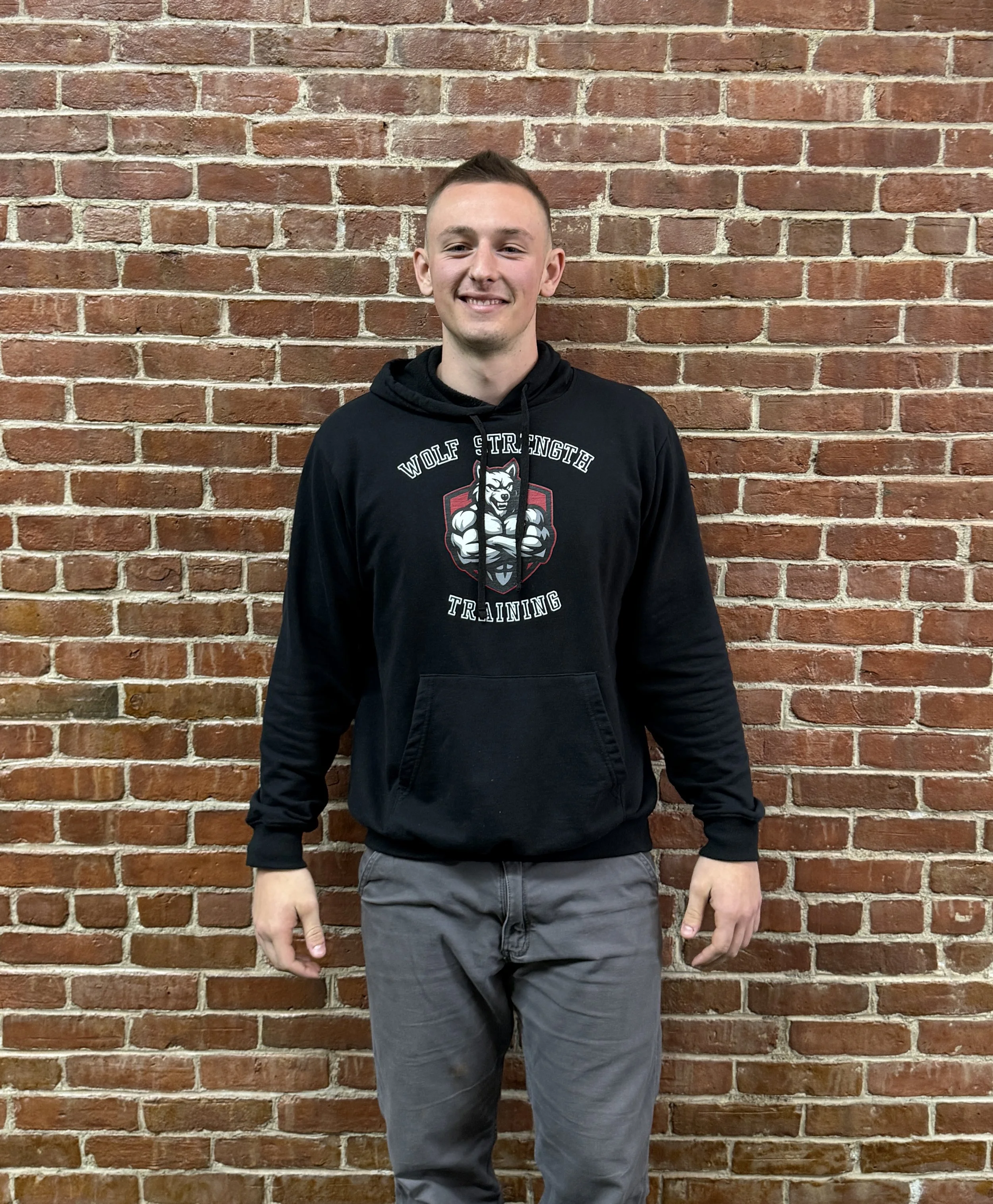 A young man with short dark hair and fair skin is standing in front of a brick wall. He is wearing a black hoodie with a wolf logo and the words 'Wolf Strength Training' printed on it, along with gray pants. He is smiling and has a casual posture with his right hand visible at his side and his left hand the same.