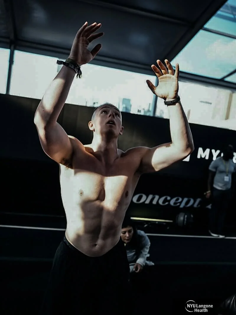 A shirtless young man with a muscular build, raising his hands above his head, appears to be at a sporting event or gym, with a woman in the background and a large window showing city buildings.