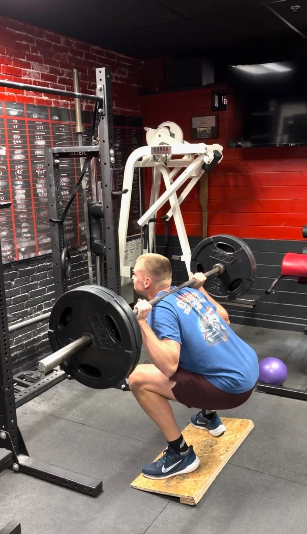 A man doing a squat exercise in a gym with a barbell loaded with weights, positioned on a wooden plank for safety.