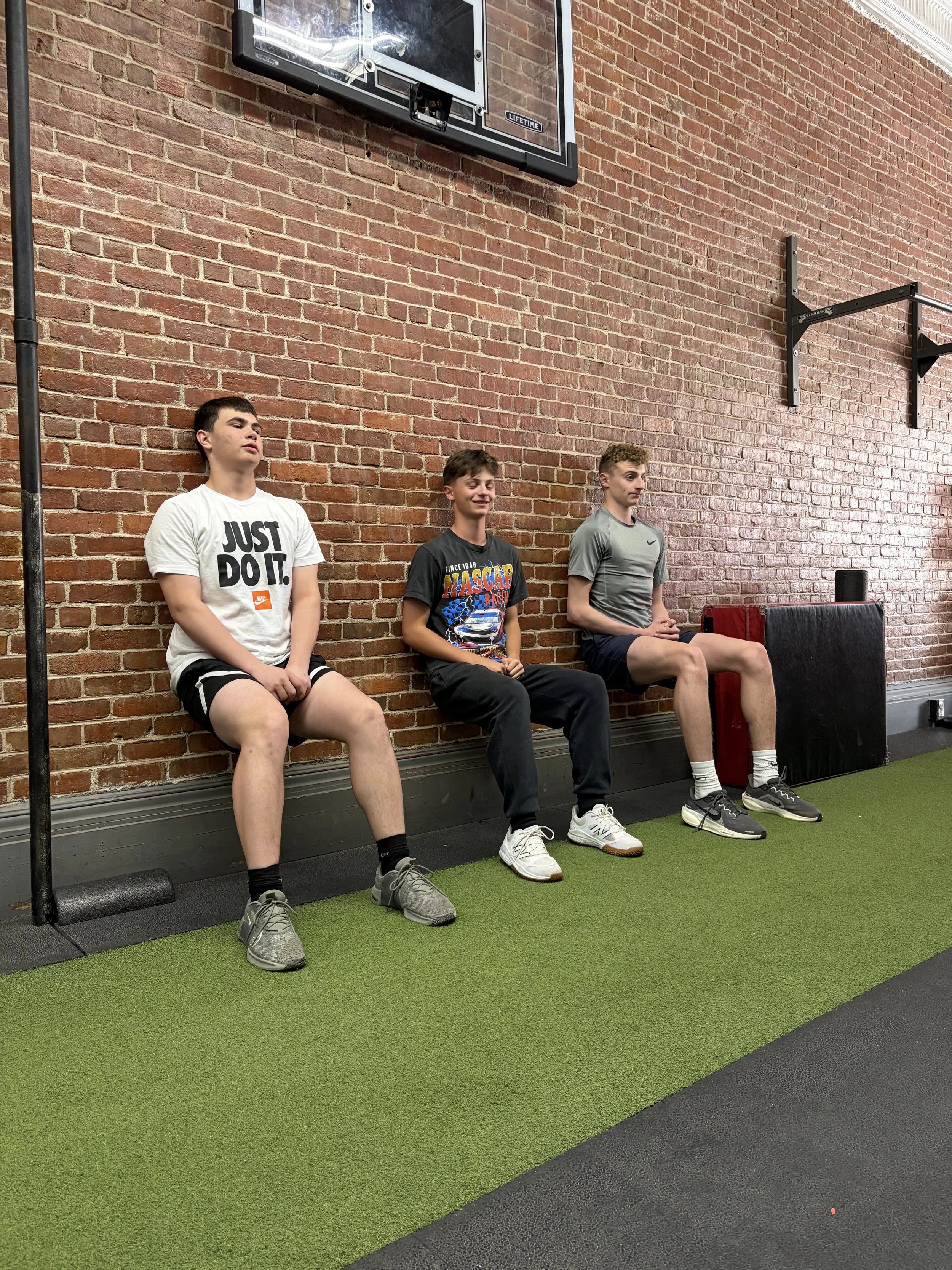 Three young men sitting against a brick wall in a gym or fitness studio. They are dressed in athletic clothes, with one wearing a white 'Just Do It' t-shirt, another in a 'NASCAR' graphic t-shirt, and the third in a gray Nike t-shirt. They are seated on a bench or ledge with a green and black rubber flooring.