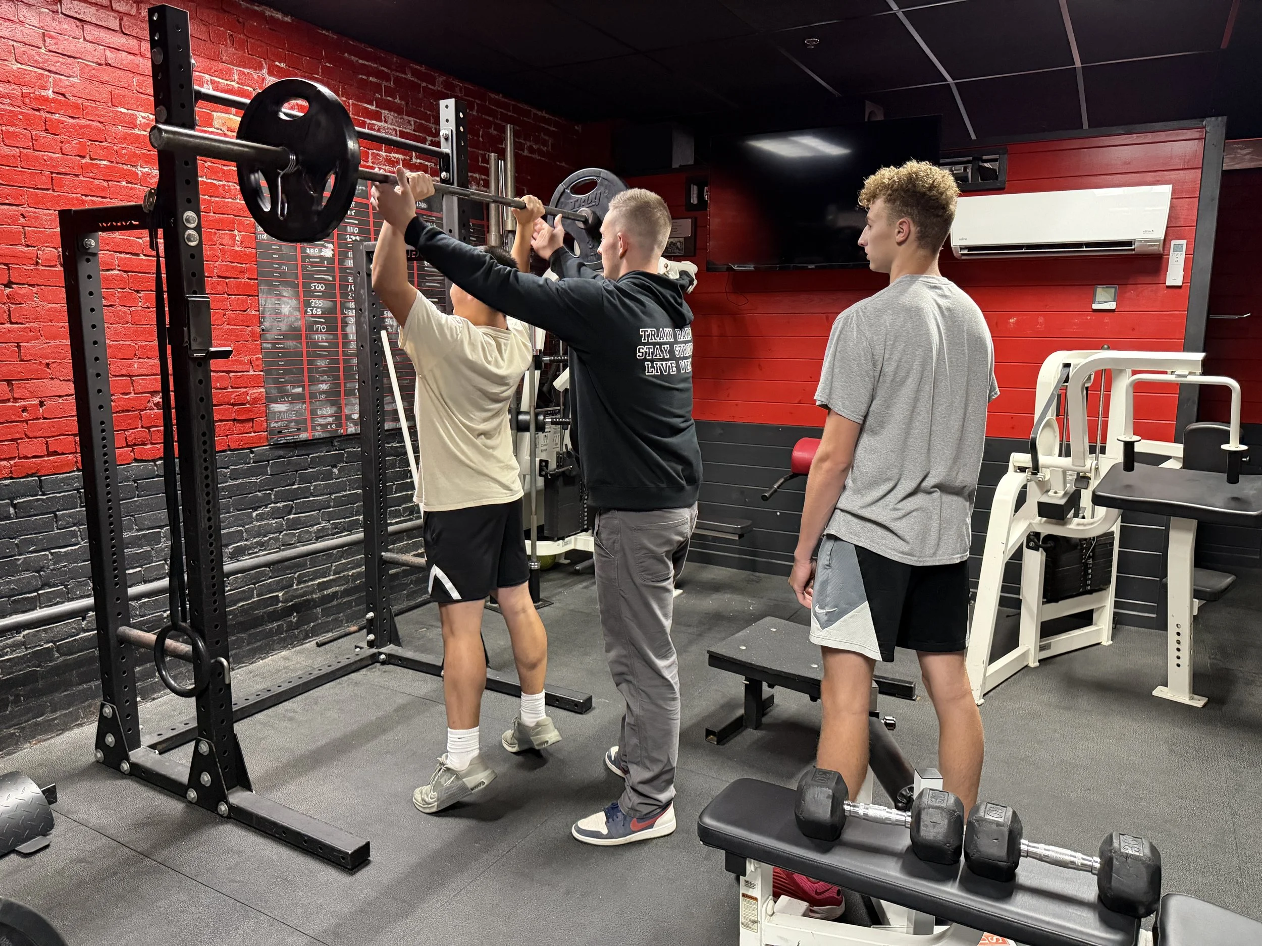 Three young men in a gym are working out, with one spotting another as he lifts a barbell with weights.