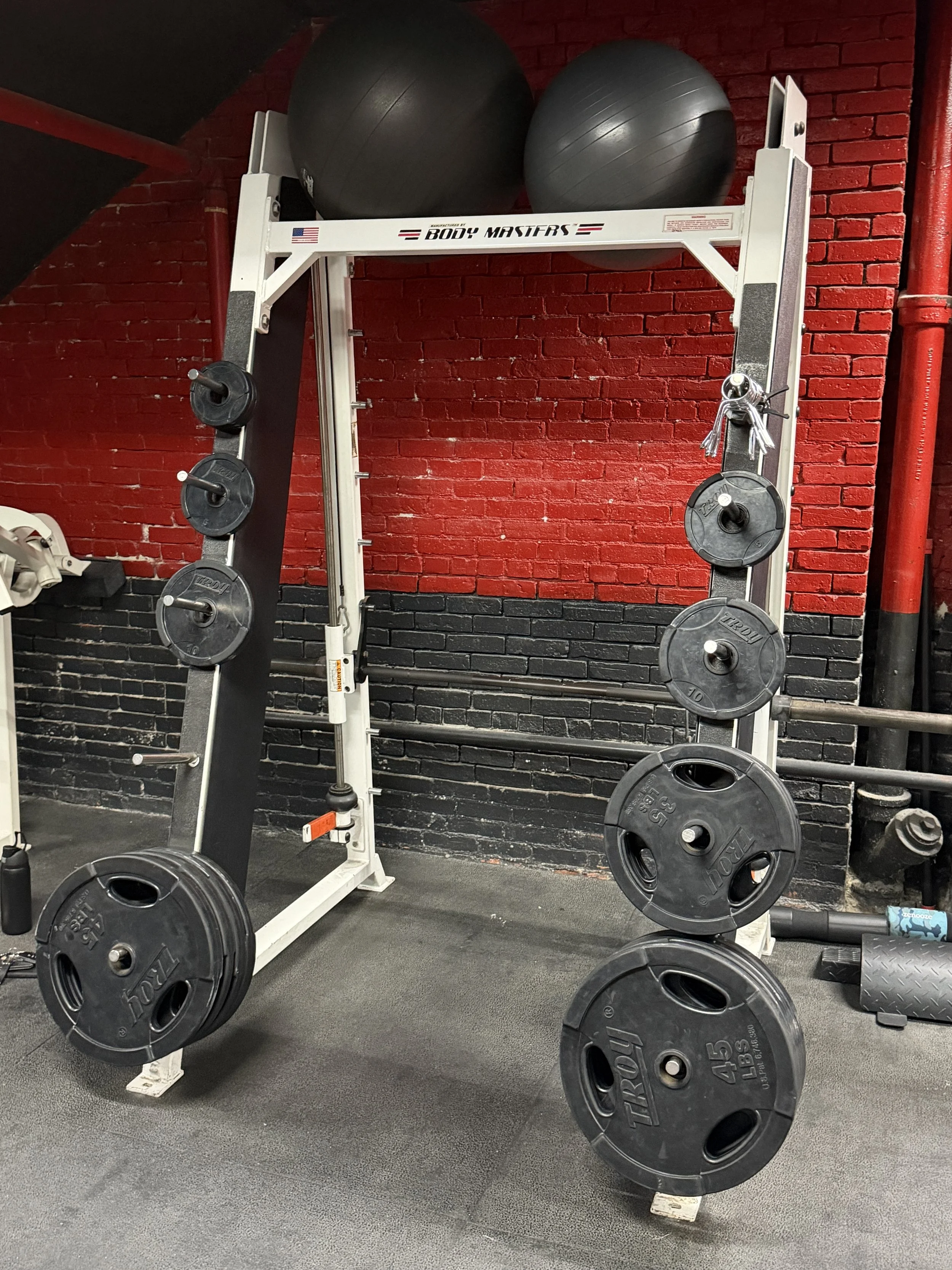 Weightlifting and gym equipment with black weight plates on a white rack, two large black exercise balls on top of a white structure, set against a red and black brick wall.
