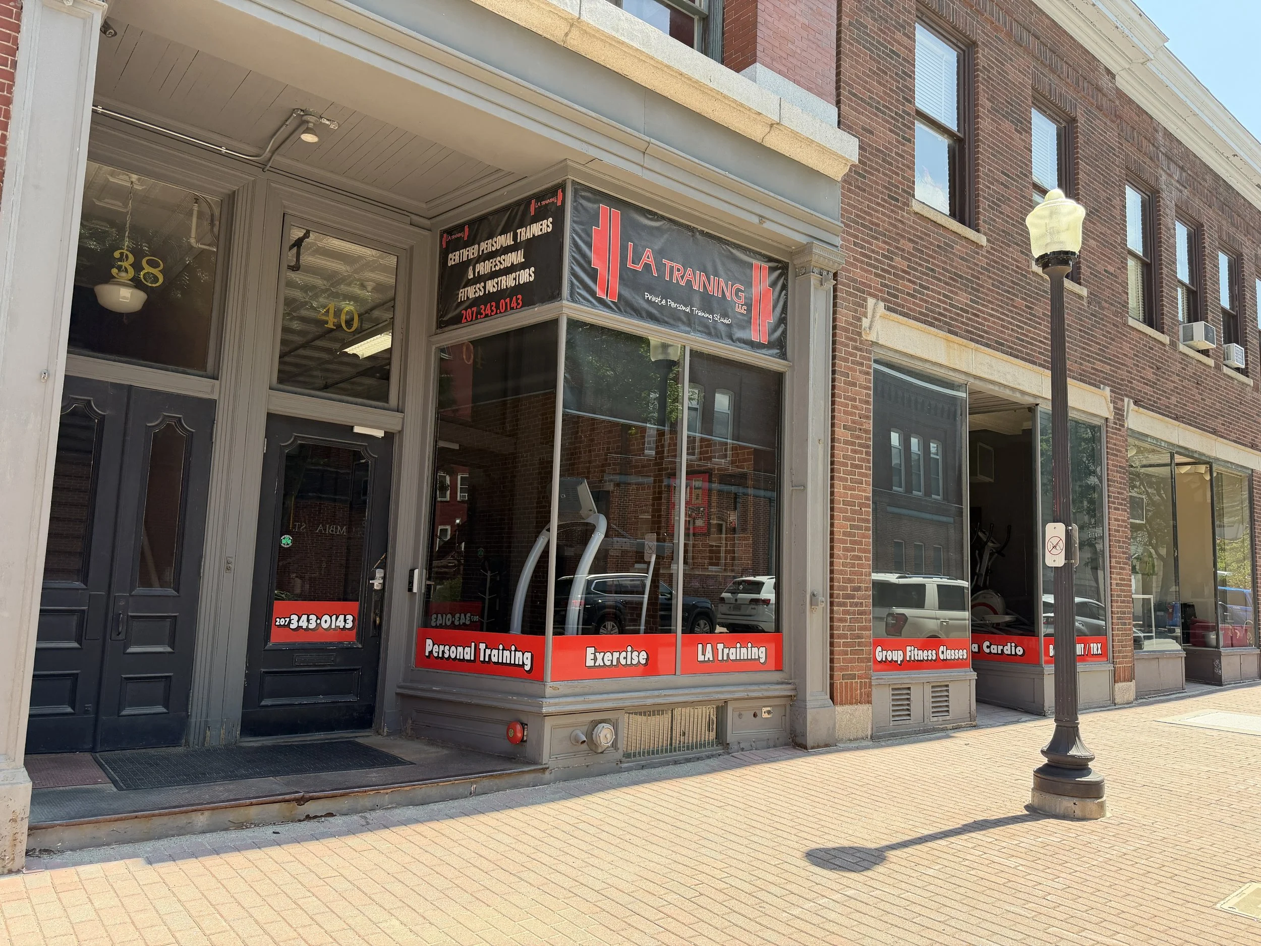 Exterior view of a brick building housing a personal training studio named LA Training, with signage indicating services like personal training, exercise, group fitness classes, and cardio. A street lamp stands on the sidewalk in front.