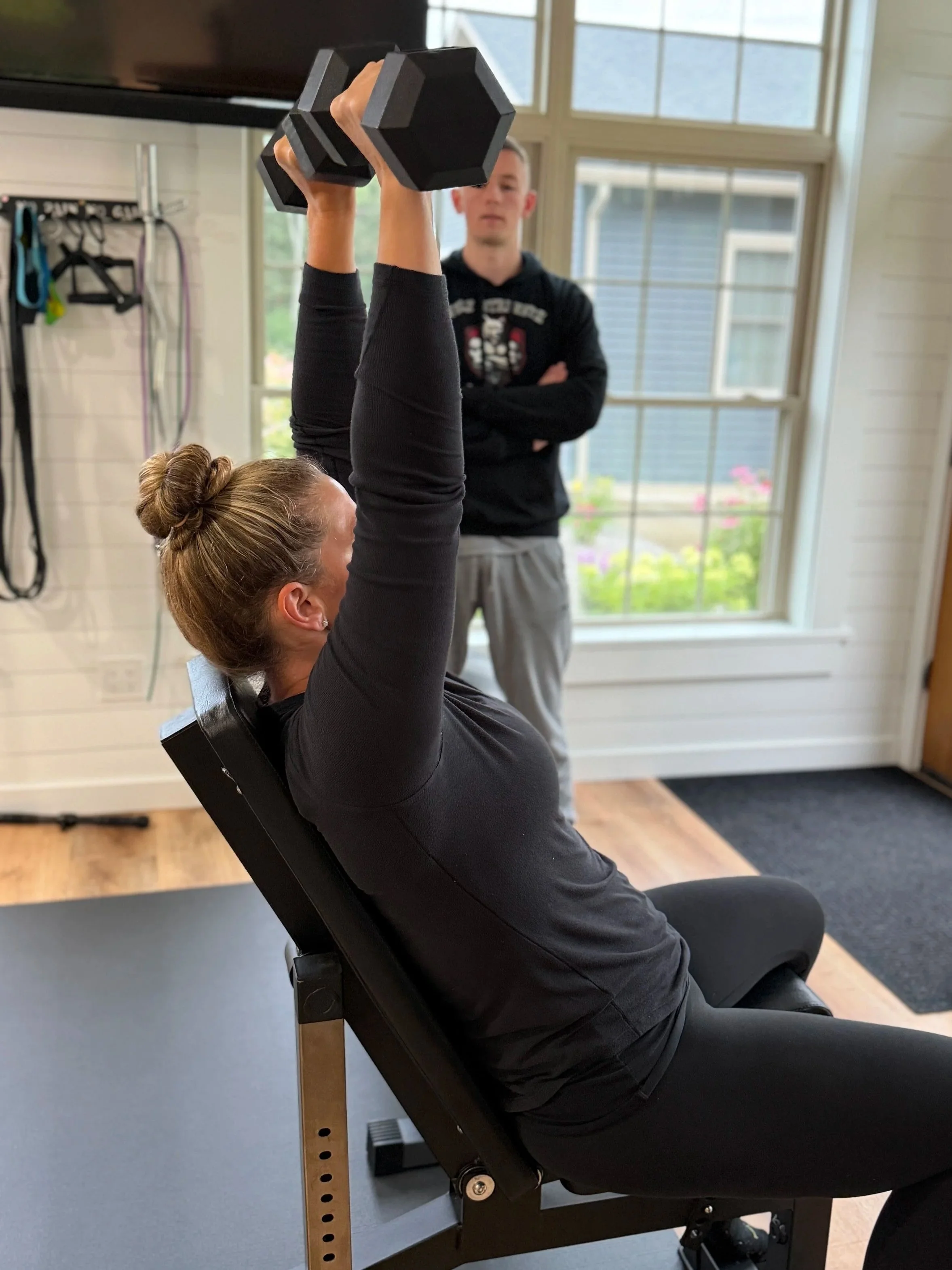 A woman is lying on an adjustable weight bench, lifting a dumbbell with her right arm while a man stands nearby observing. The woman has her hair in a bun and is wearing a dark long-sleeve shirt, and the man is in a black hoodie with arms crossed.