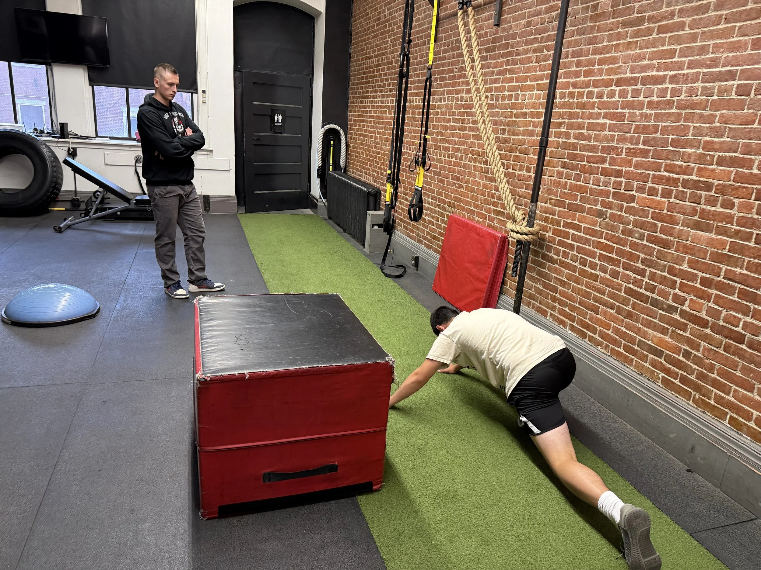 Two men are exercising in a gym with a brick wall. One man is standing with arms crossed, the other is doing push-ups with hands on the ground. Gym equipment such as a large tire, pull-up bars, resistance bands, and a red padded box are visible.