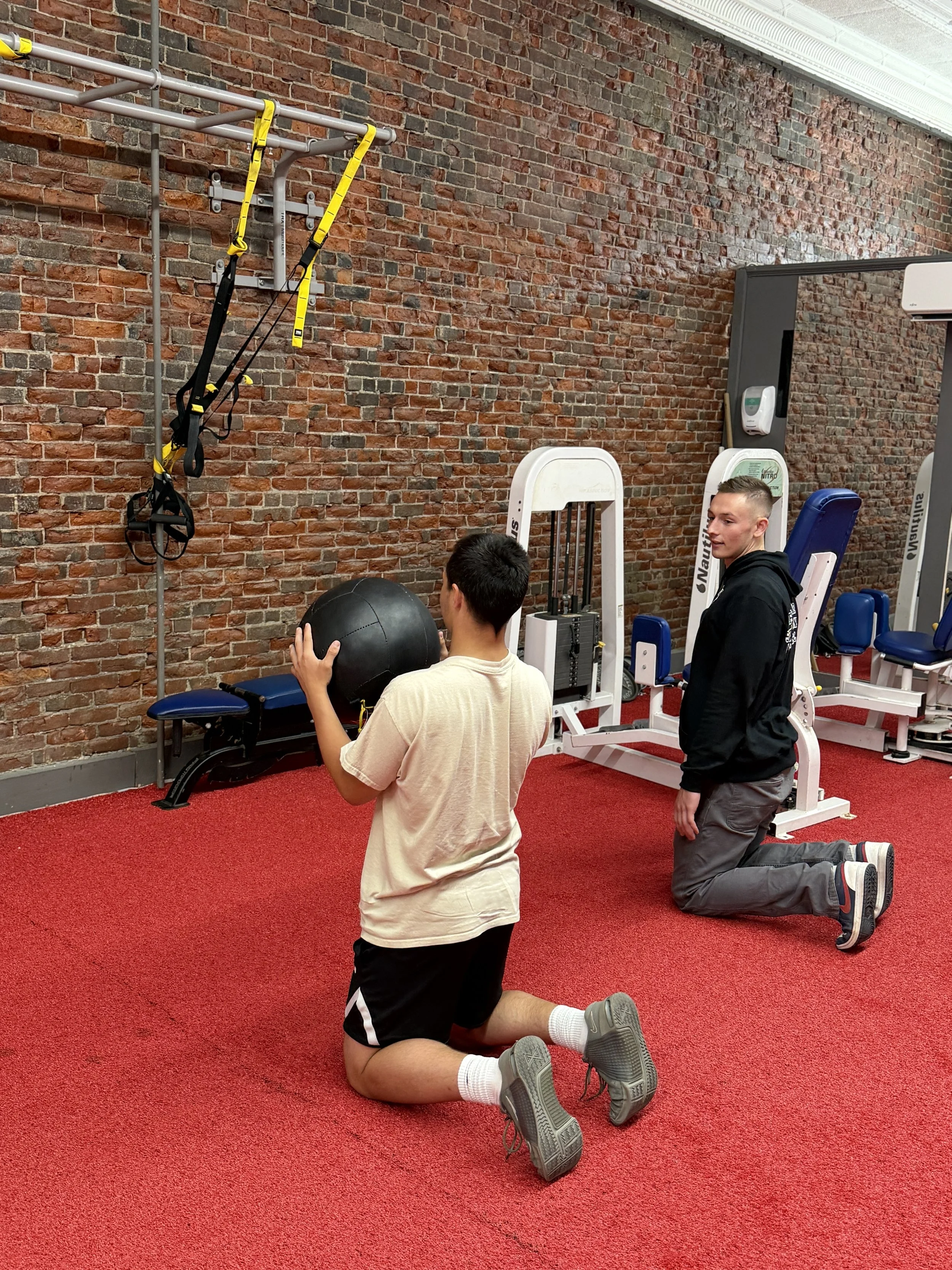 Two young men exercising in a gym with exposed brick walls. One is kneeling on the red carpeted floor, holding a medicine ball, while the other is standing nearby, looking at the kneeling man, with gym equipment around them.