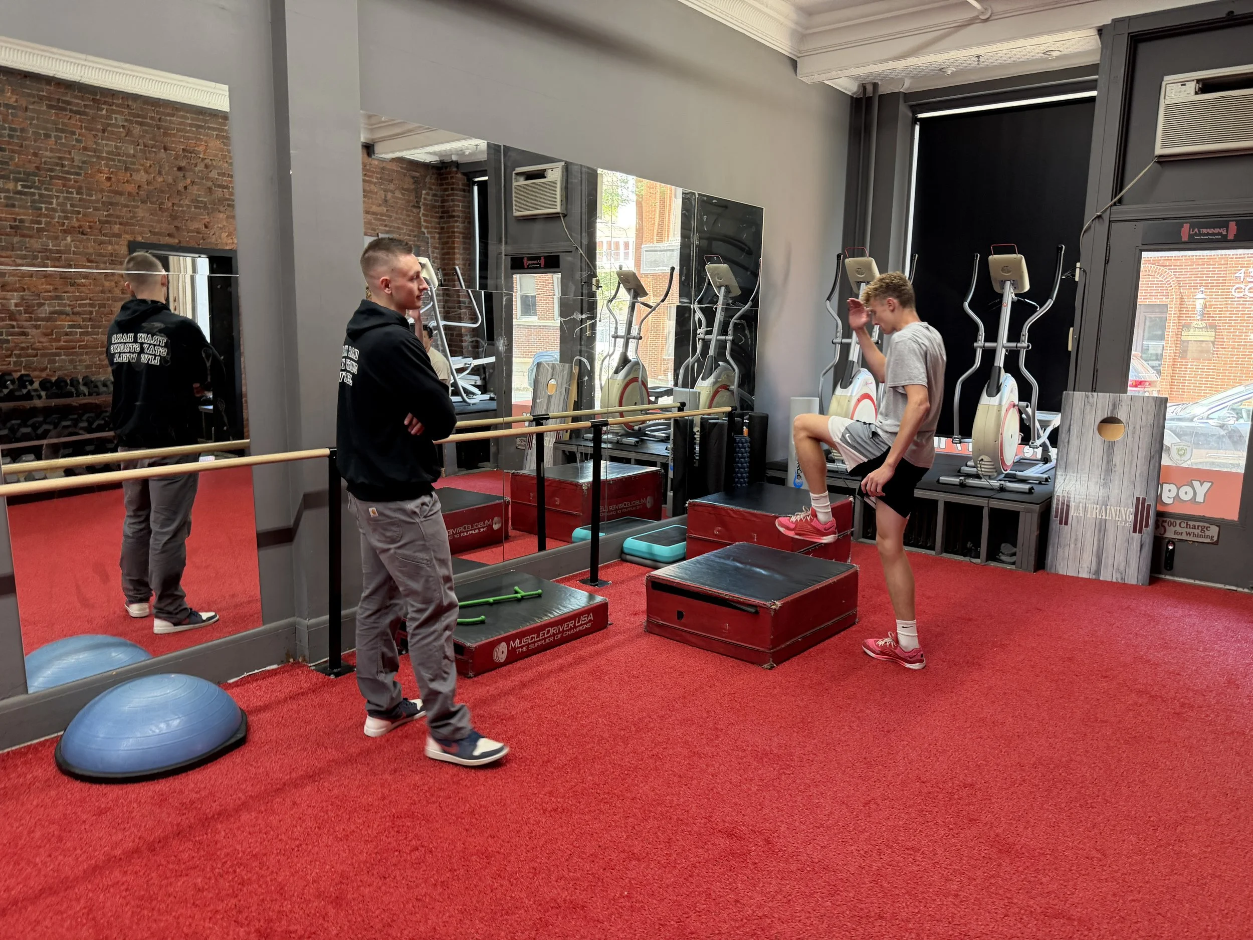 Young man stepping onto a red platform in a gym with two older men watching, surrounded by exercise equipment, mirrors, and brick walls.