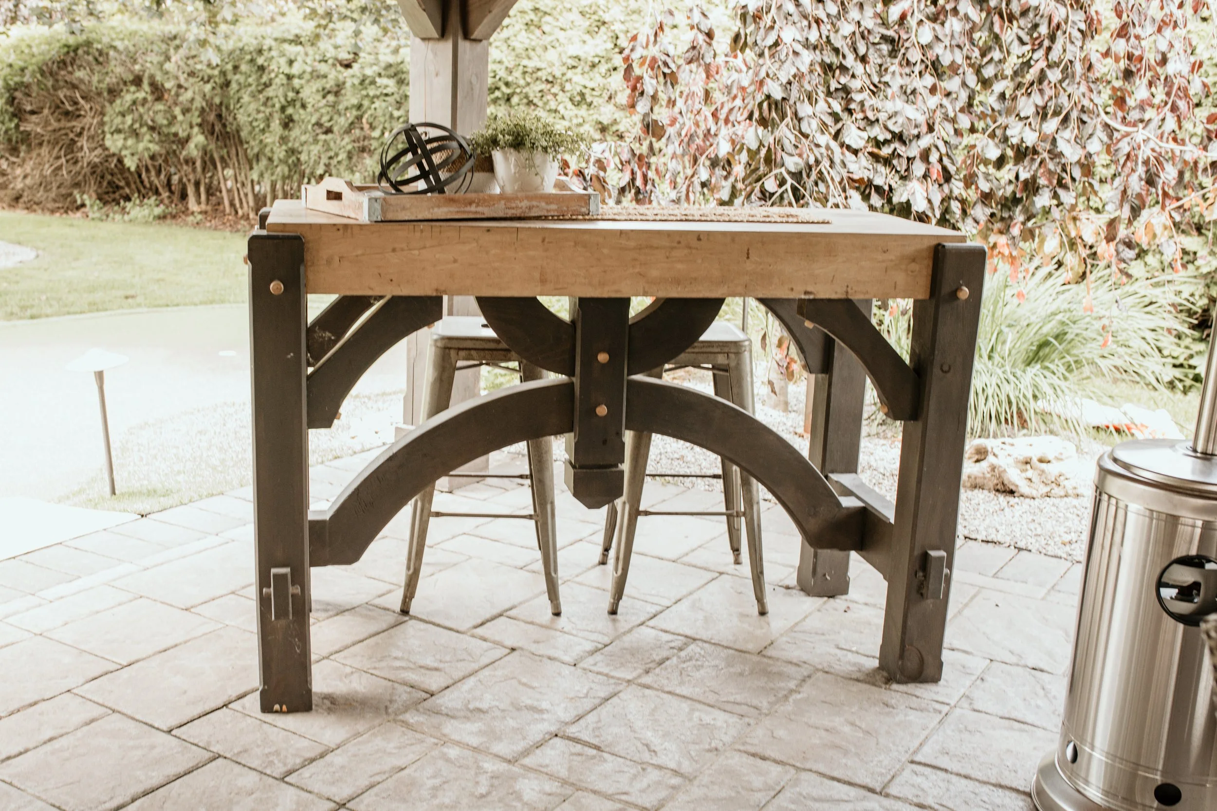 Outdoor patio scene featuring a wooden table with black metal supports, a potted plant, decorative objects, and classical chairs with metal legs, set against a background of bushes and trees.