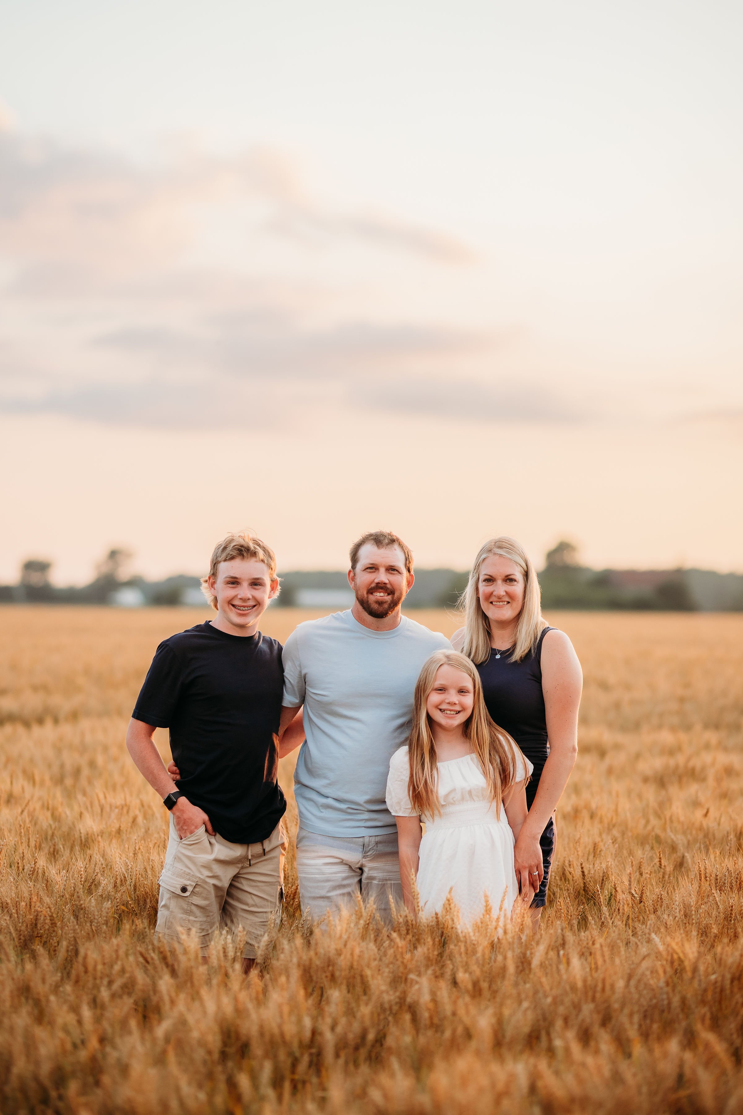 A family of four standing in a golden field during sunset, smiling and posing for a photo.