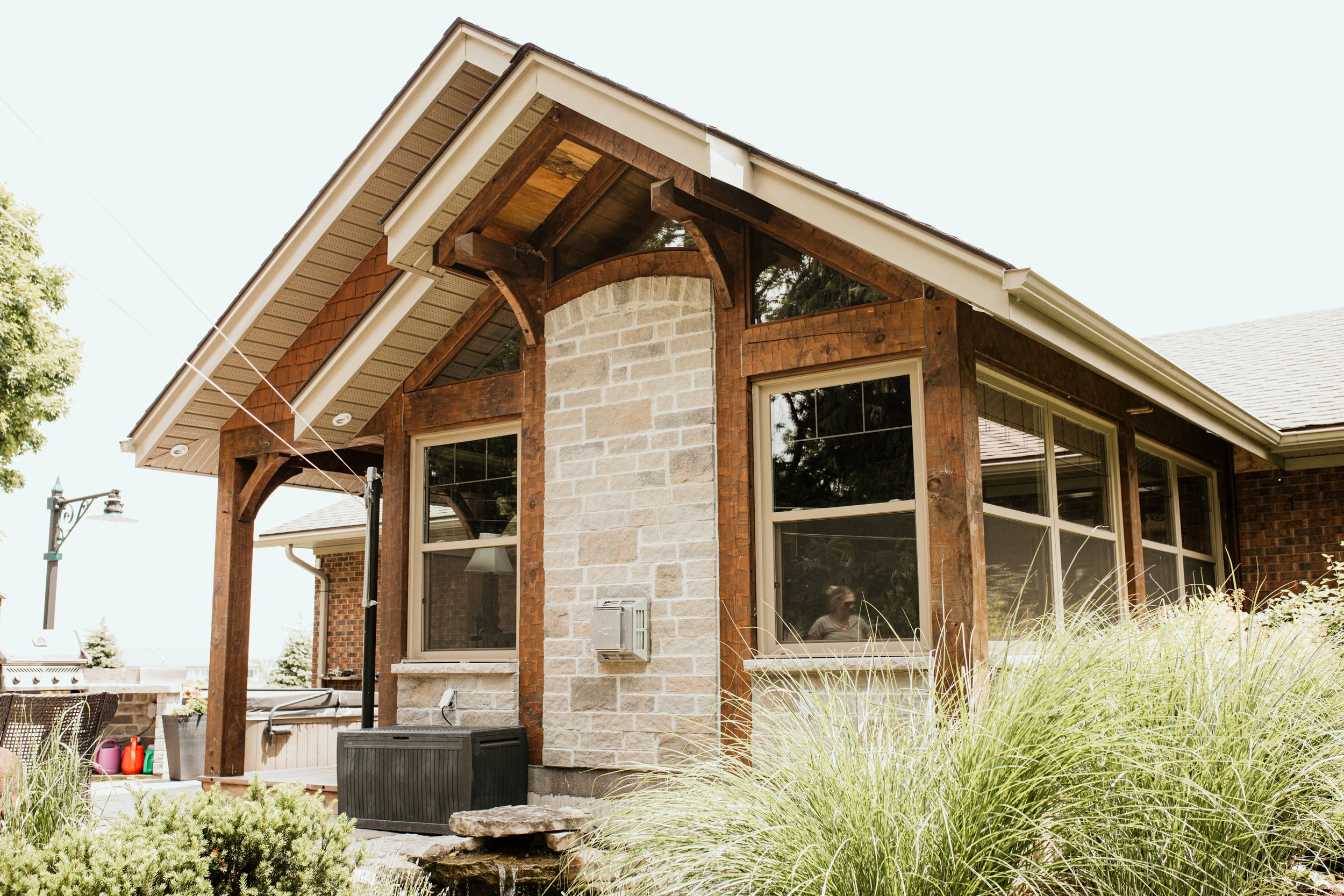 Close-up of a house exterior with brick and stone walls, wooden framing, and large windows, with a garden and outdoor furniture.