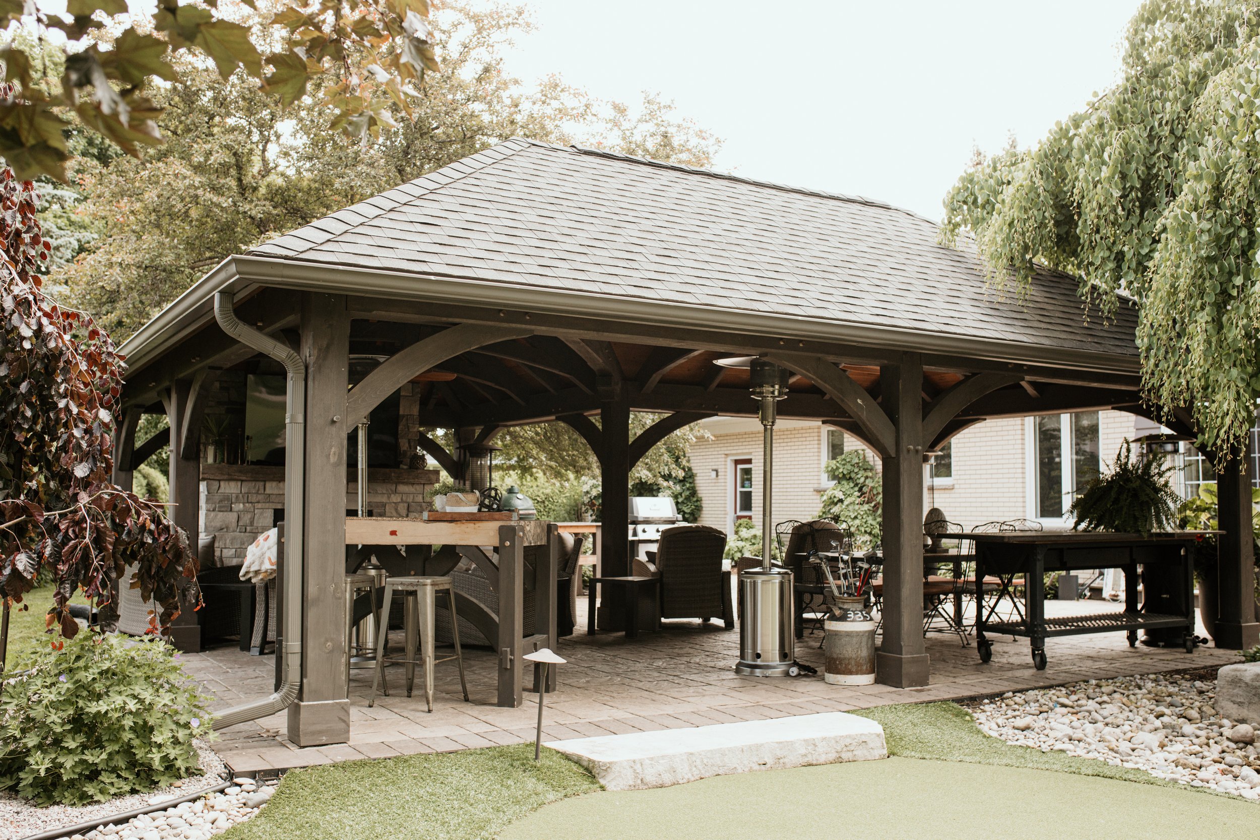 Outdoor patio with a wooden pavilion, seating, and cooking area, surrounded by trees and landscaping.