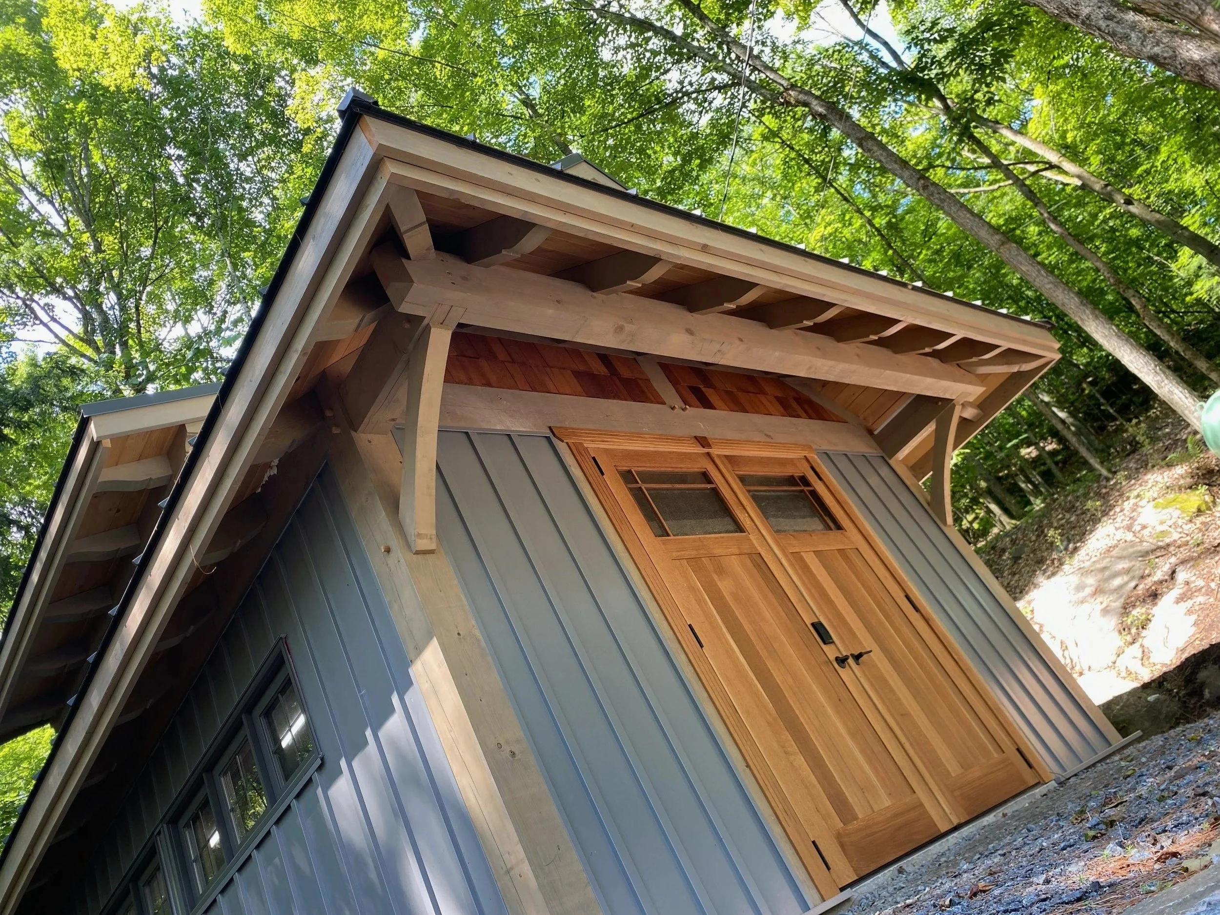 A house under construction with wooden siding, metal panels, and an open roof showing roof framing and trees around.