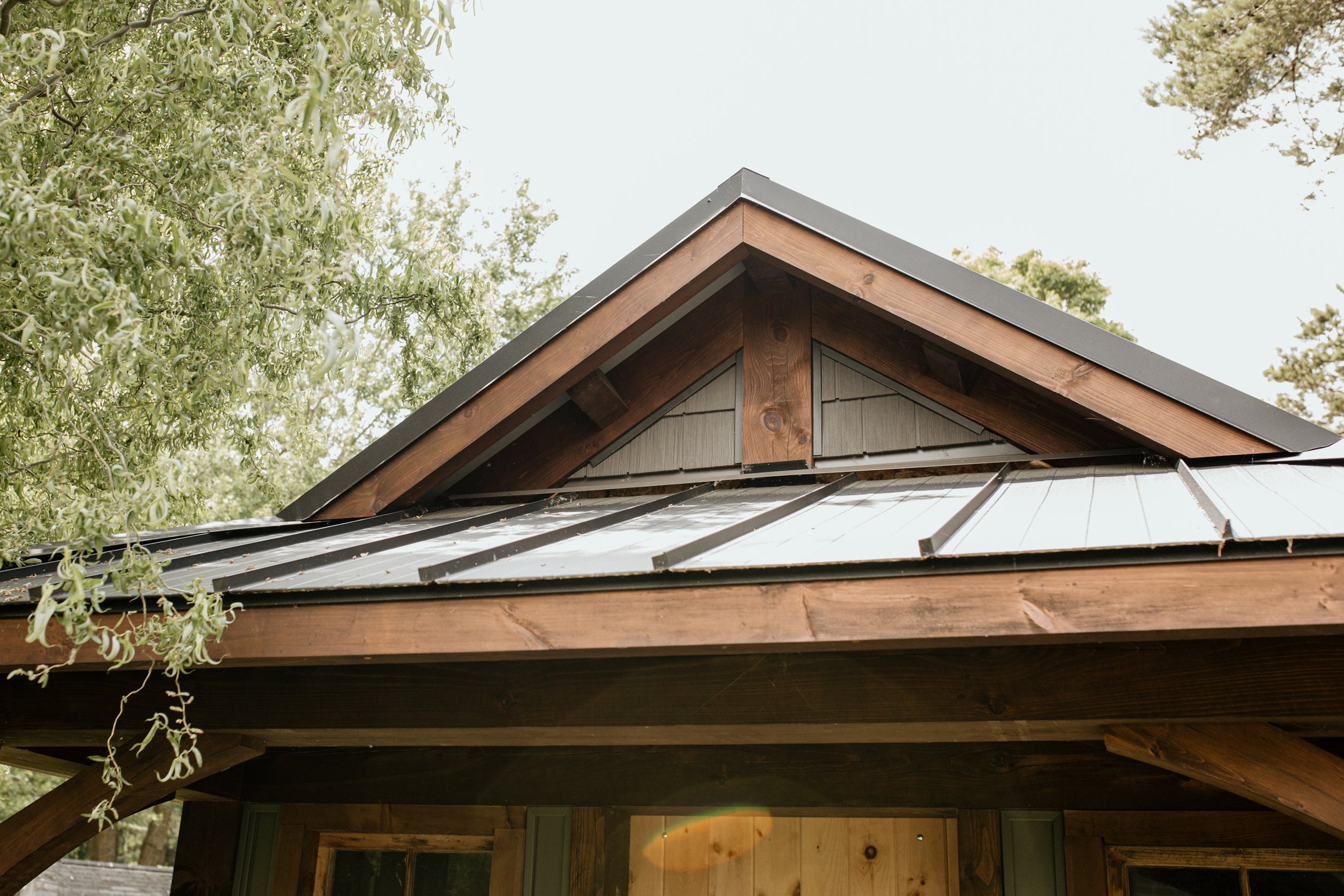 Close-up of a house roof with metal and wooden details, surrounded by tree branches with green leaves