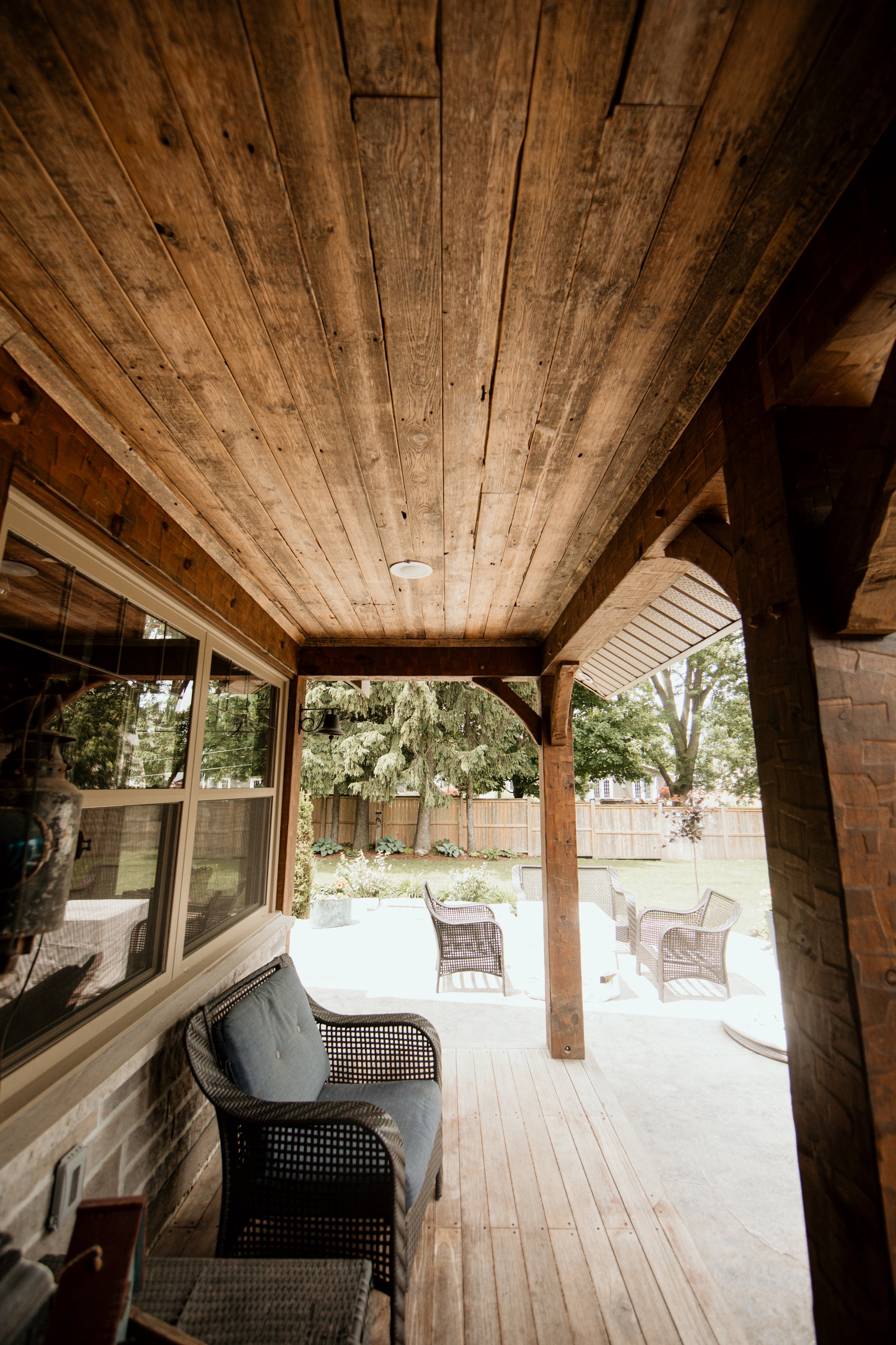 Covered porch with wooden ceiling, wicker furniture, and a backyard with trees, a wooden fence, and patio chairs.