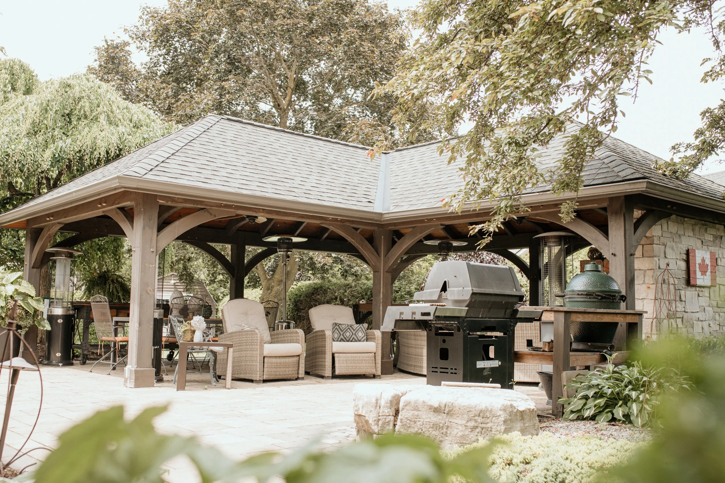Outdoor patio with seating area, a barbecue grill, and outdoor heating lamps under a wooden pavilion surrounded by trees and plants.