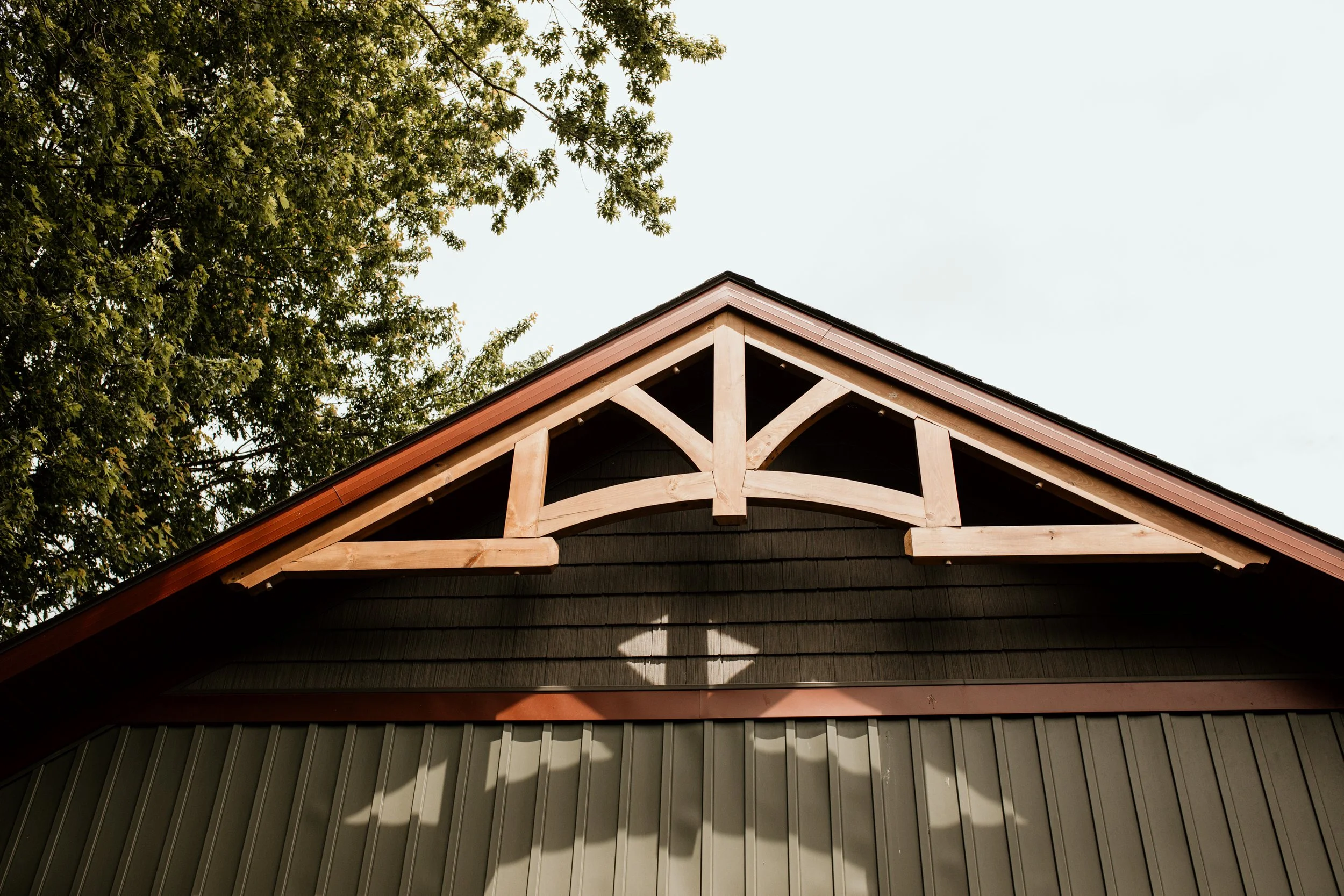 Close-up of the gable end of a house with dark siding, showing decorative wooden trusses at the peak of the roof and a tree in the background.