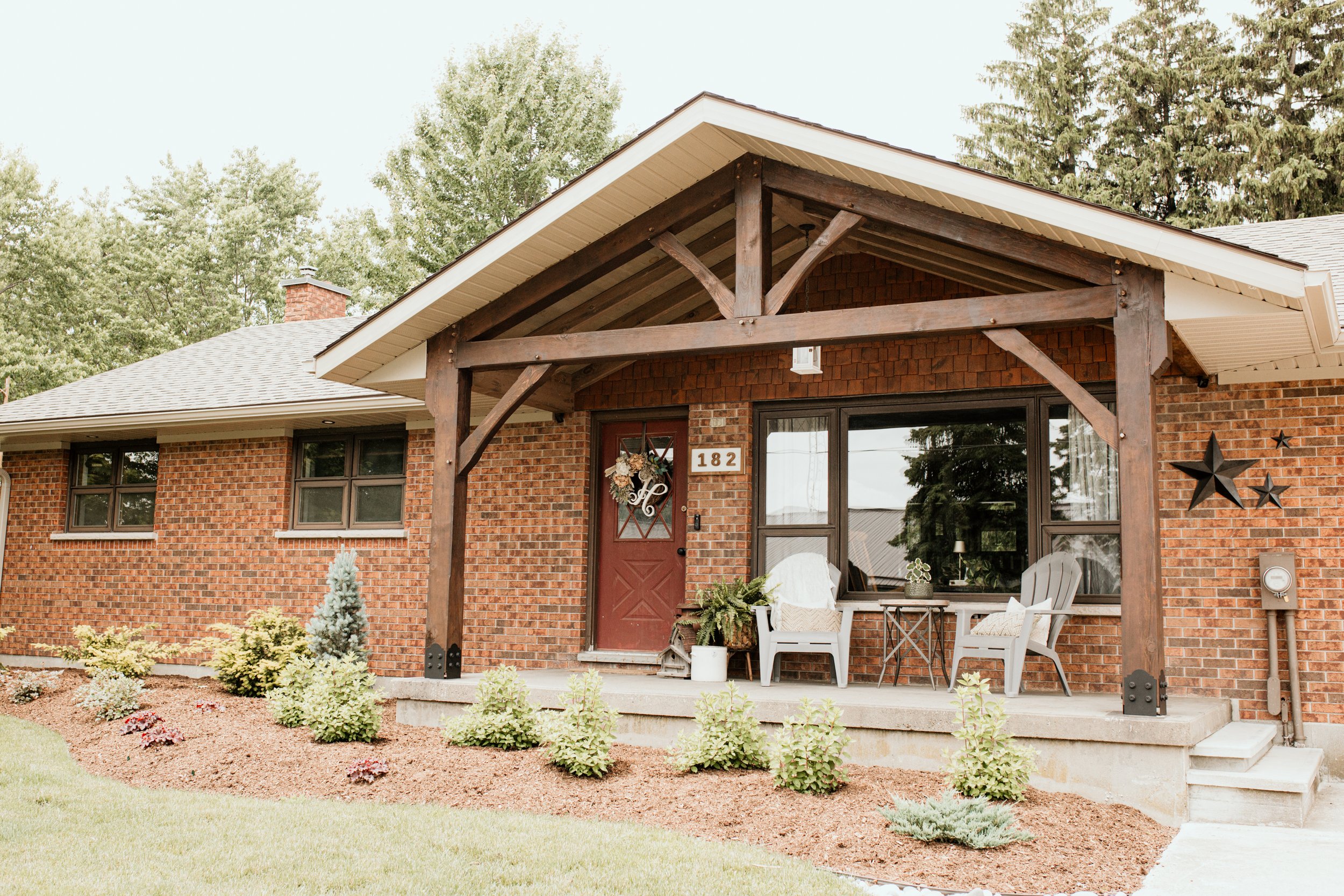 Front porch of a brick house with a wooden gabled roof, featuring two white porch chairs, potted plants, and decorative star wall art, with a lawn and trees in the background.