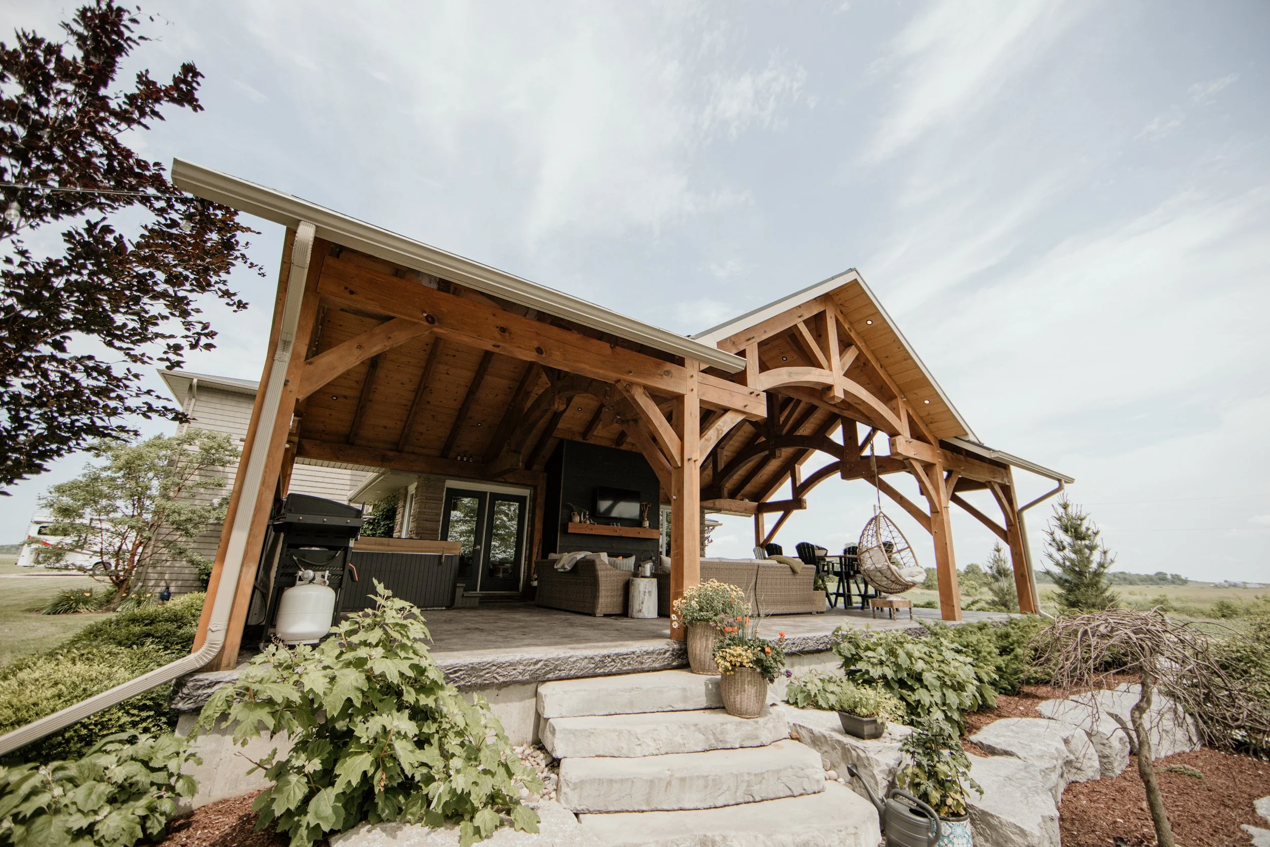 A covered outdoor patio with wooden beams, outdoor furniture, potted plants, a hanging chair, and a landscaped garden with shrubs and trees.