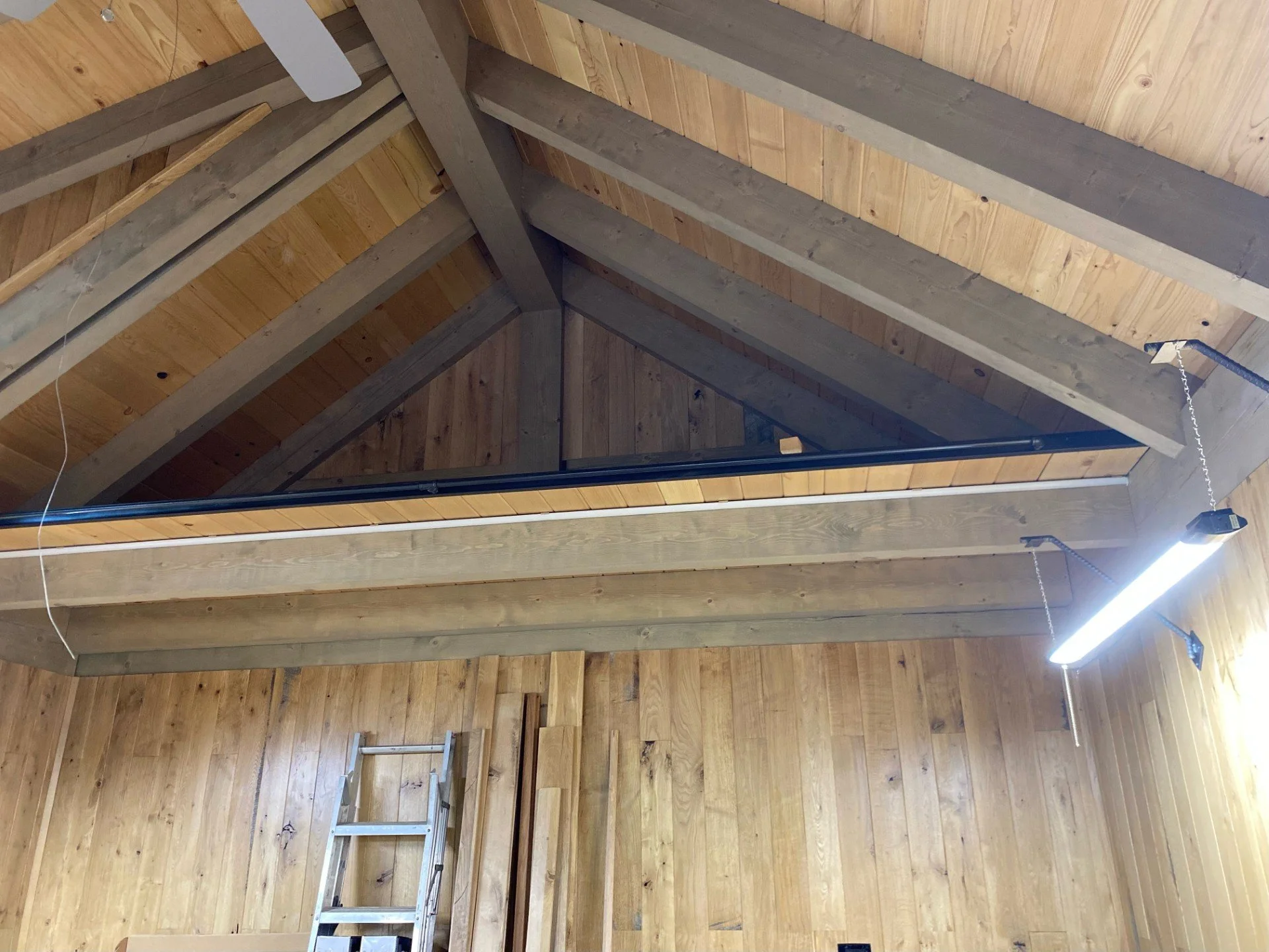 Interior of a wooden building with exposed ceiling beams, a hanging fluorescent light, and a metal ladder leaning against the wood-paneled wall.