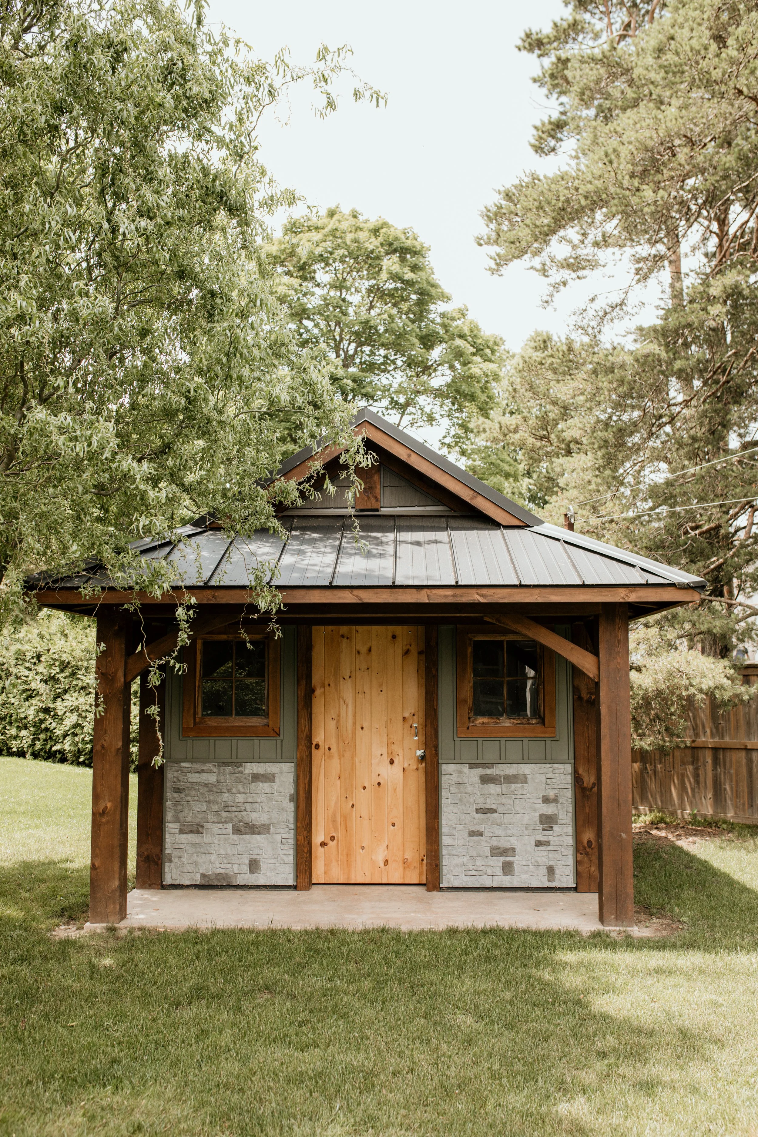 Small wooden shed with stone and wood exterior, metal roof, and small windows, surrounded by green grass and trees.