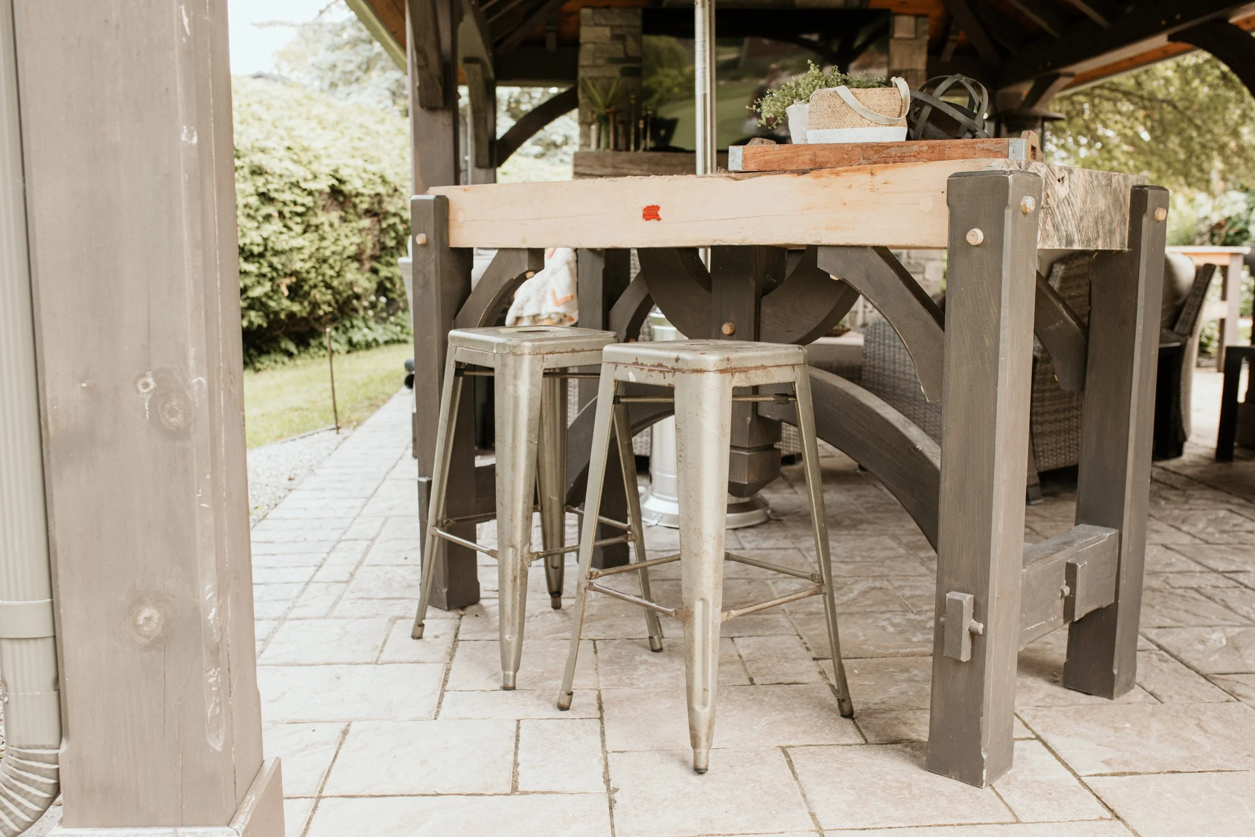 Outdoor patio area with a wooden table, two metal barstools, and a stone tile floor. The table has decorative items on top, and there is greenery in the background under a covered porch.