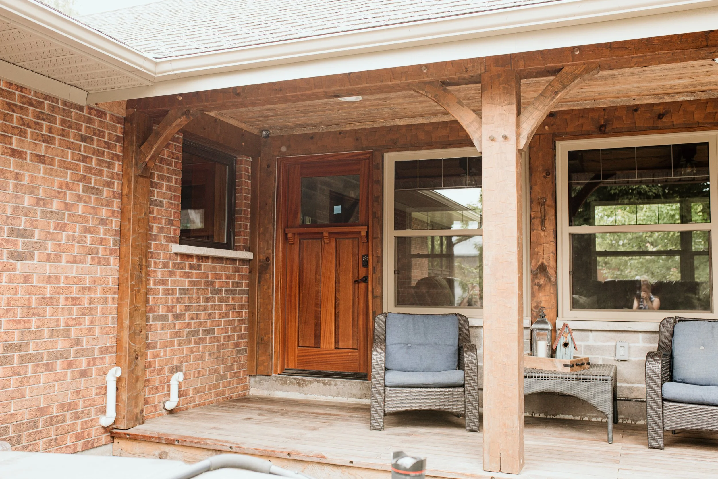 Front porch area with brick wall, wooden door, two large windows, outdoor wicker chairs with blue cushions, small table with decorative items, and wooden beams supporting the roof.