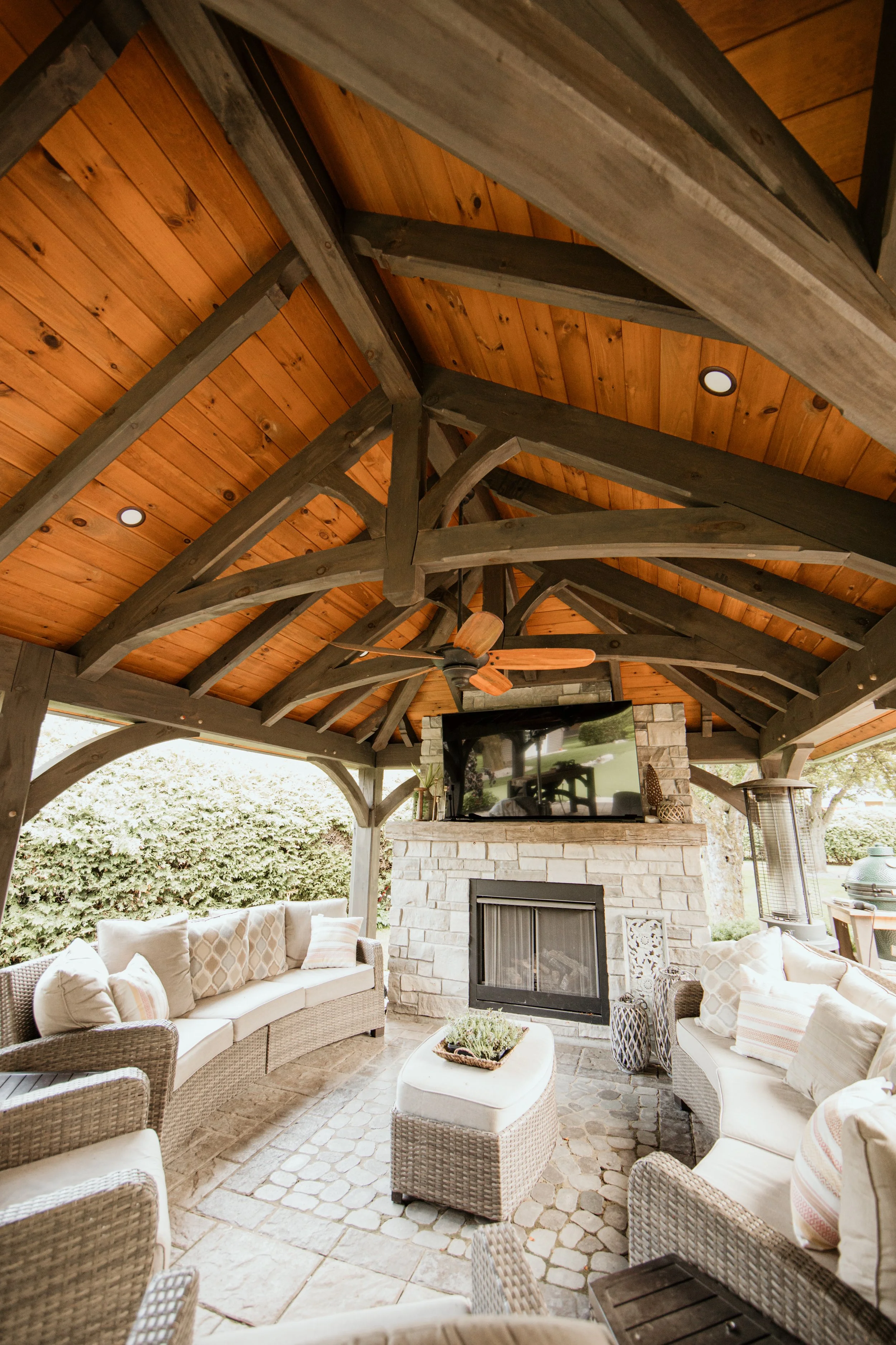 Outdoor patio seating area with wicker sofas, cushions, a stone fireplace, and a flat-screen TV under a wooden ceiling with exposed beams.