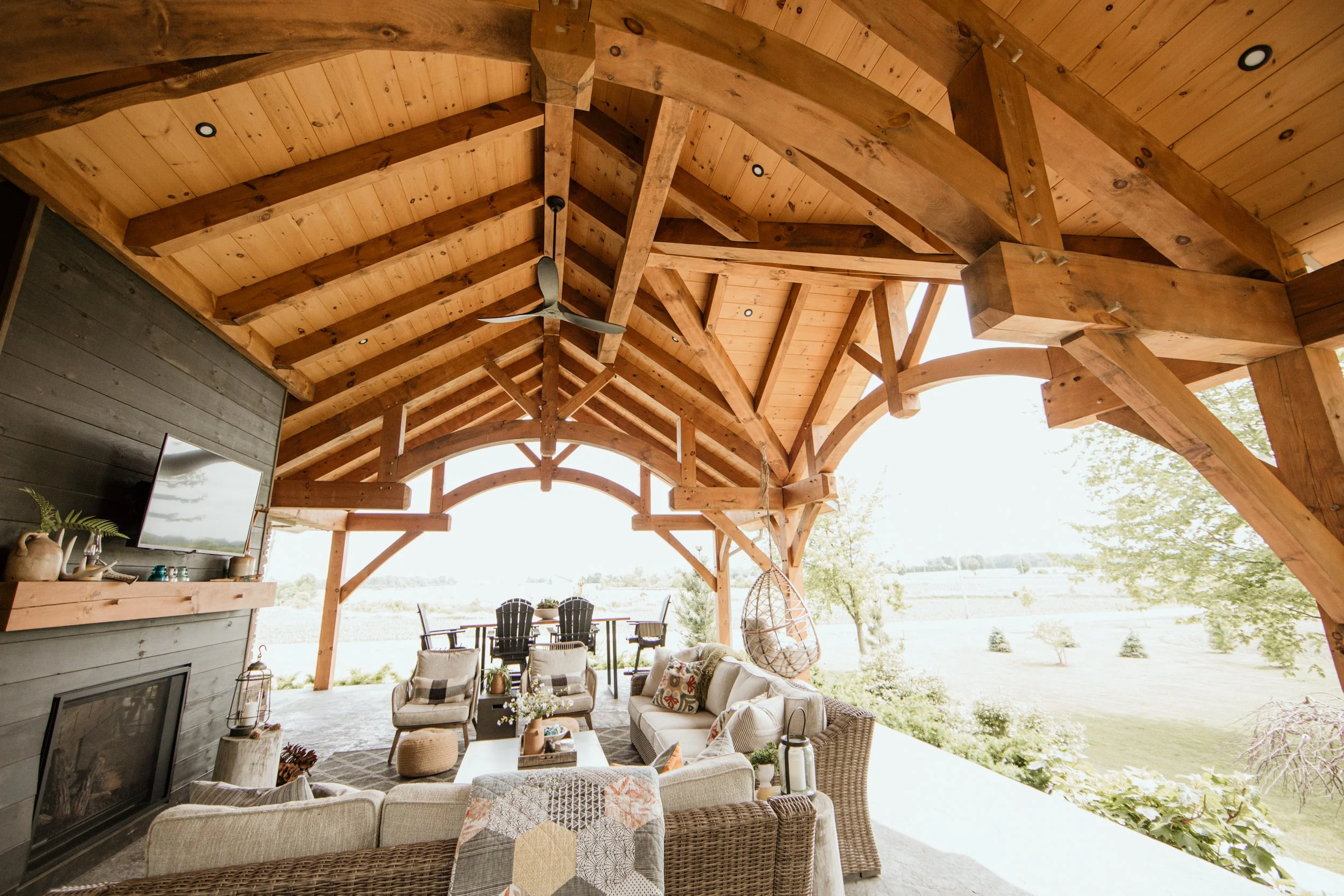 Covered outdoor patio with wooden beams and ceiling, outdoor furniture including wicker and cushioned chairs, table with flowers, hanging egg chair, and a mountain landscape view with trees in the background.