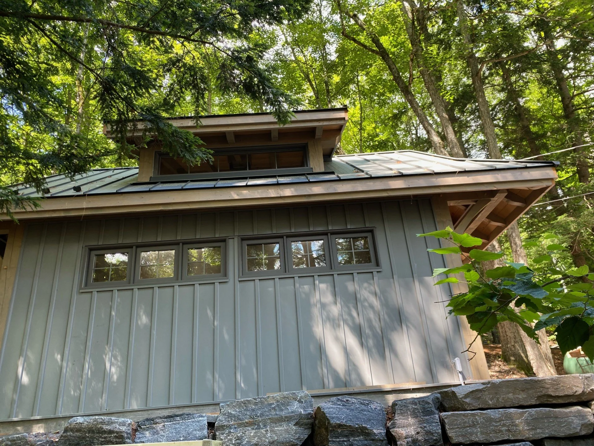 A house in a wooded area with a metal roof and gray vertical siding, surrounded by trees and green foliage.