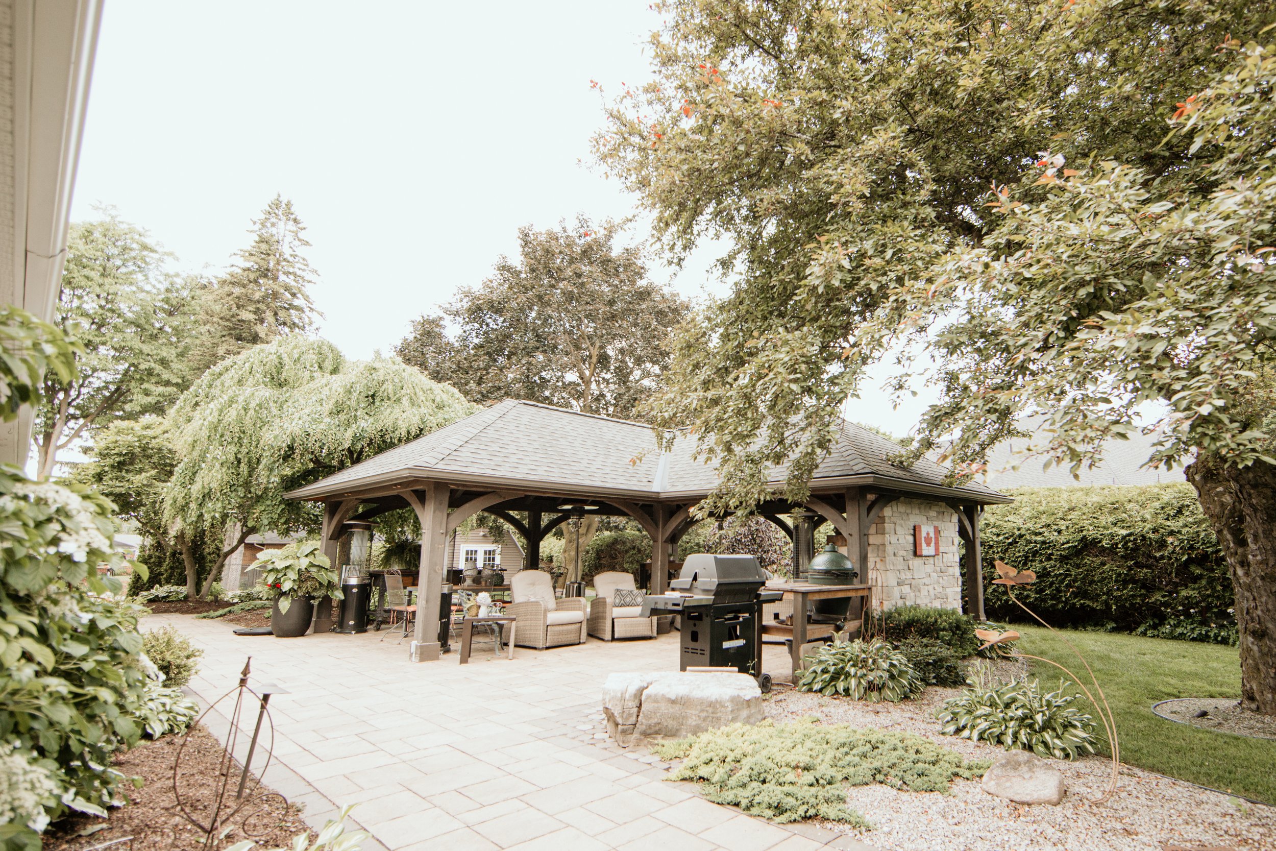 Backyard patio with a pavilion, outdoor furniture, a grill, and lush green trees and plants.