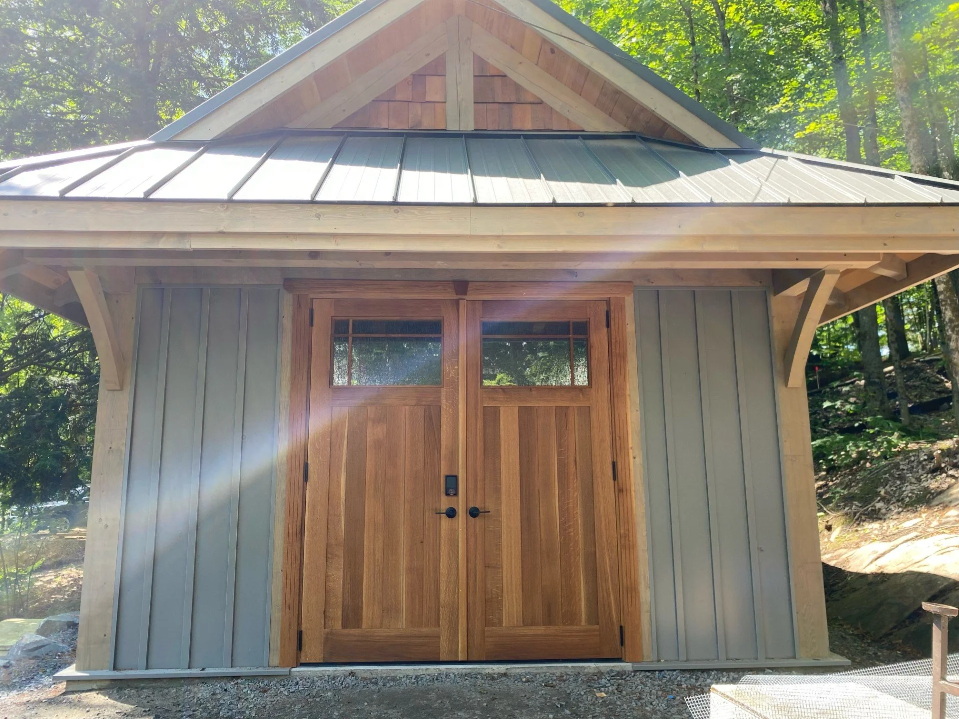 Small wooden building with double wood doors, metal roof, and surrounding trees, in a wooded area.