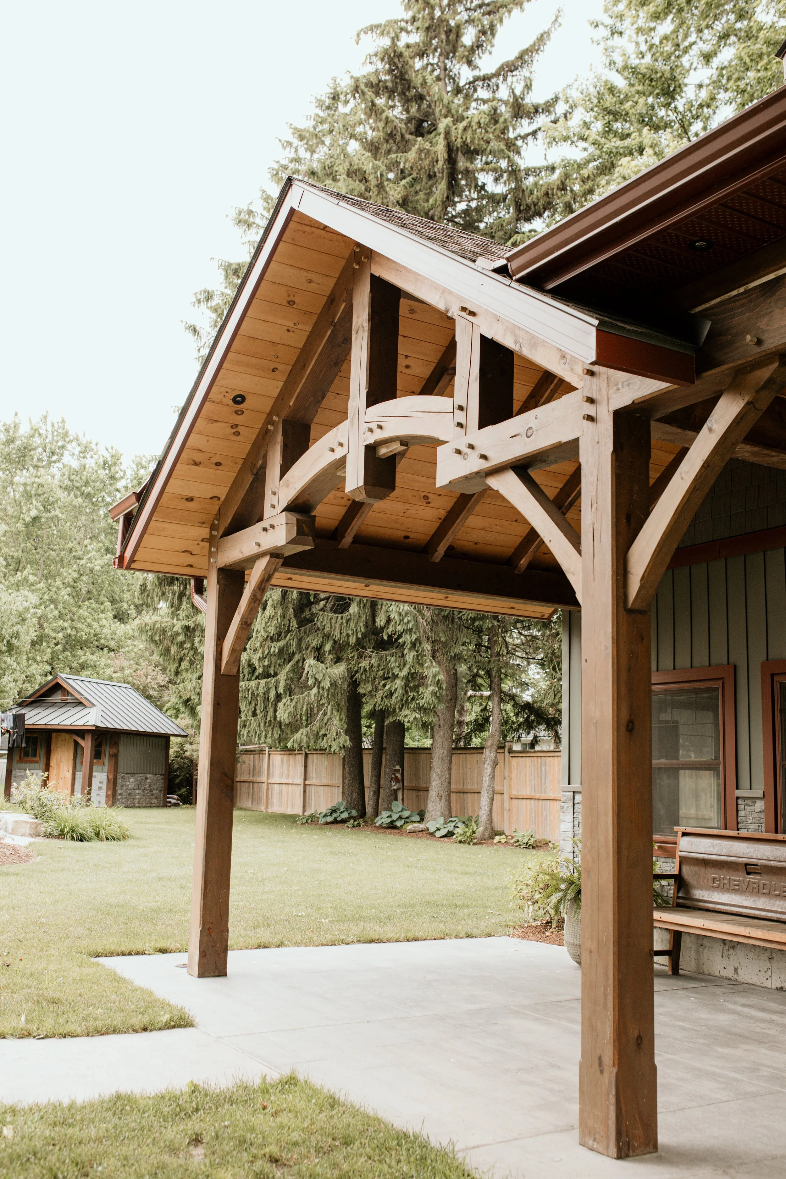Part of a house with a wooden porch structure supported by wooden beams, a concrete patio floor, a green yard with trees, and a small shed in the background.