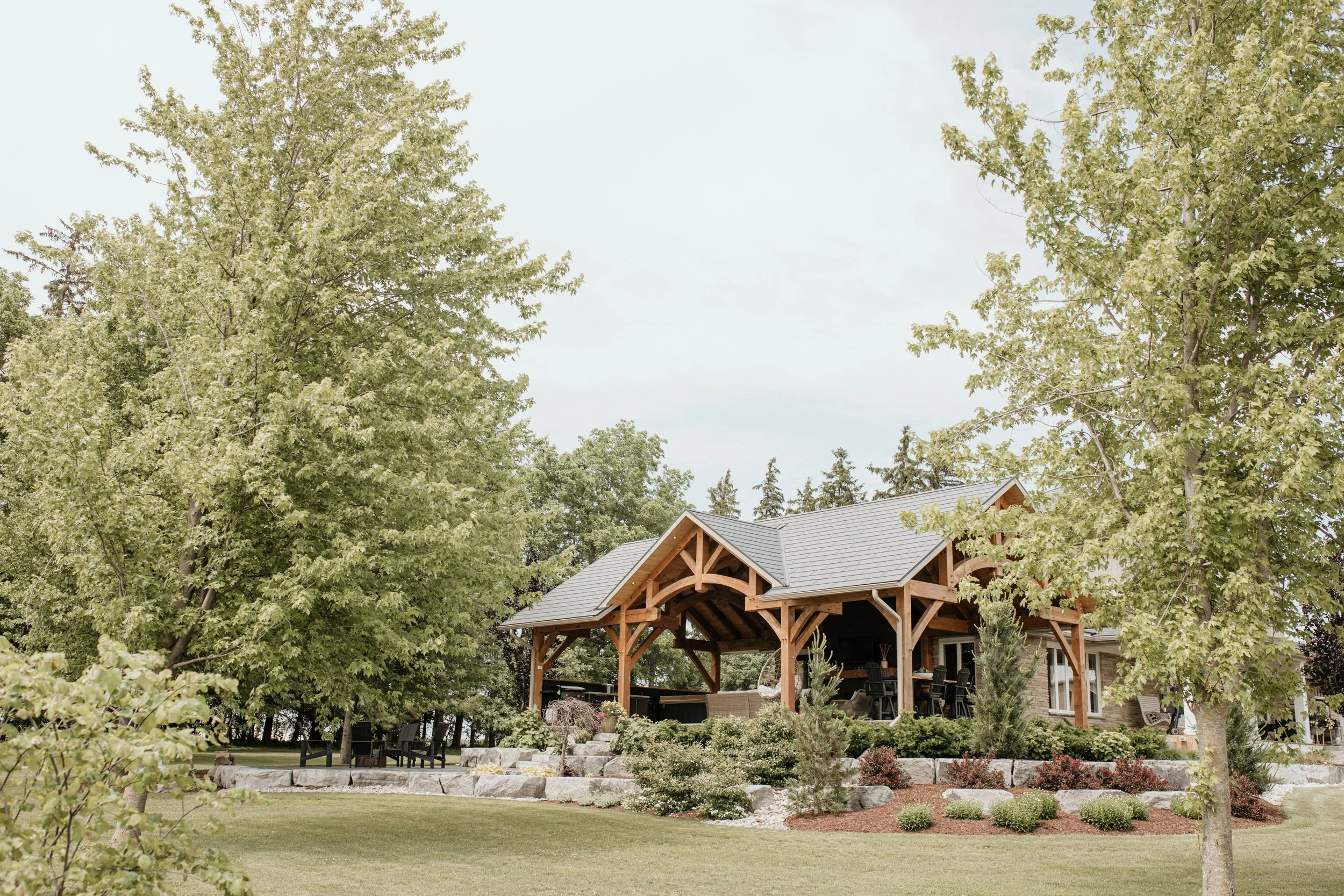 A large house with a wooden porch and a gray roof, surrounded by trees and landscaped yard with bushes and plants.