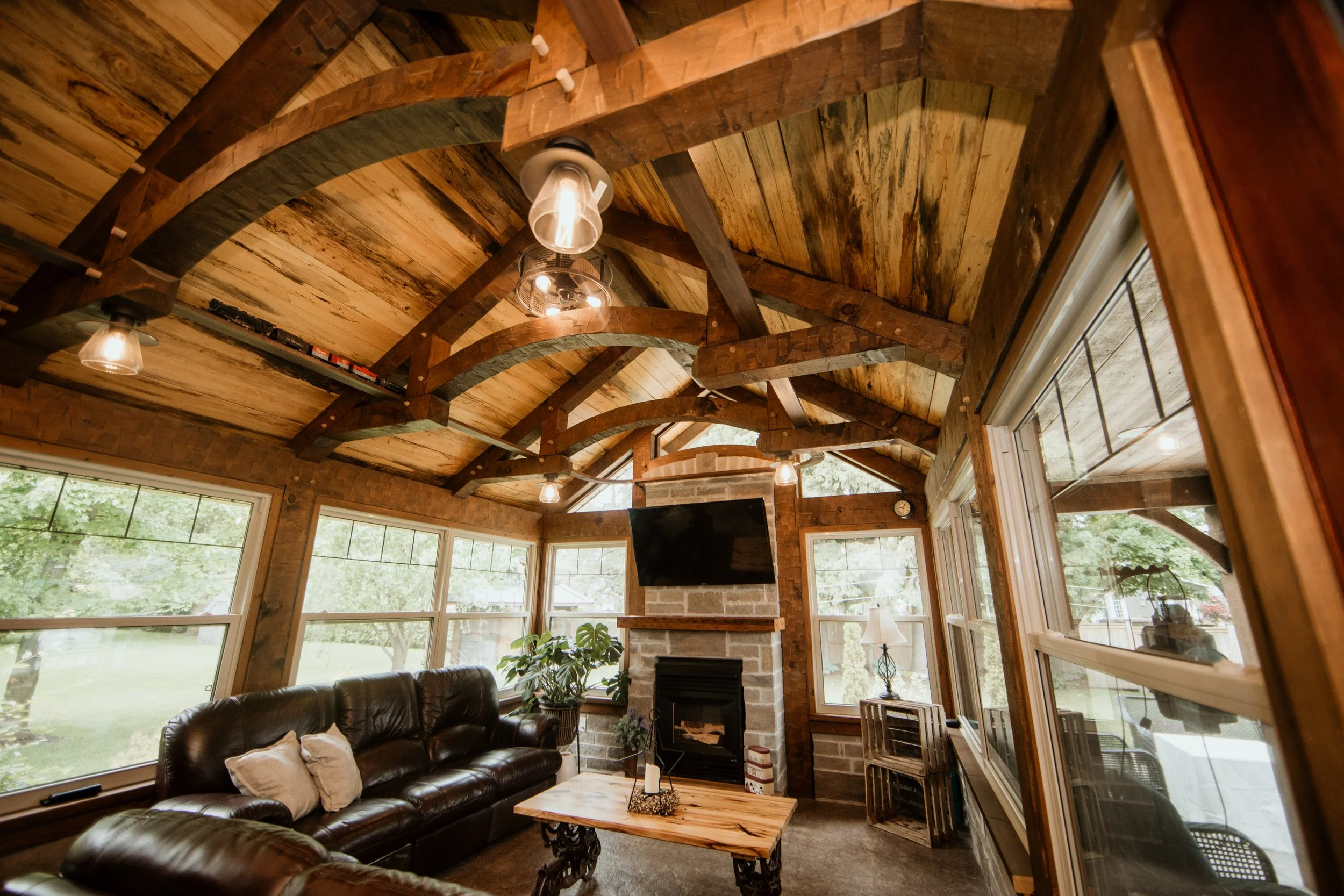 Living room with large windows, a stone fireplace with a mounted TV, brown leather sofas, a wooden coffee table, and rustic wooden ceiling beams.