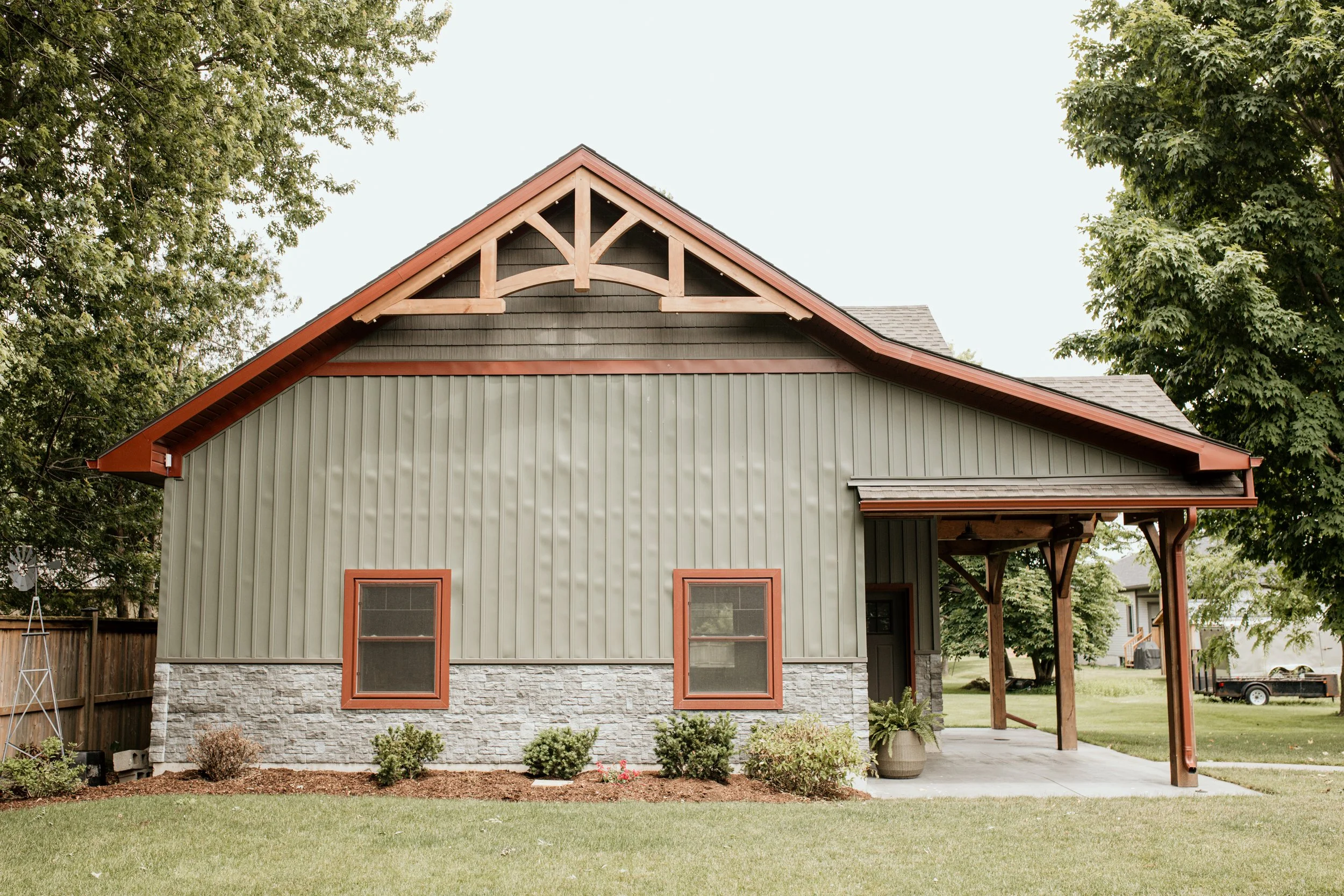 Front view of a house with a green exterior, stone base, two small windows with brown trim, and a porch supported by wooden posts, surrounded by a lawn and trees.