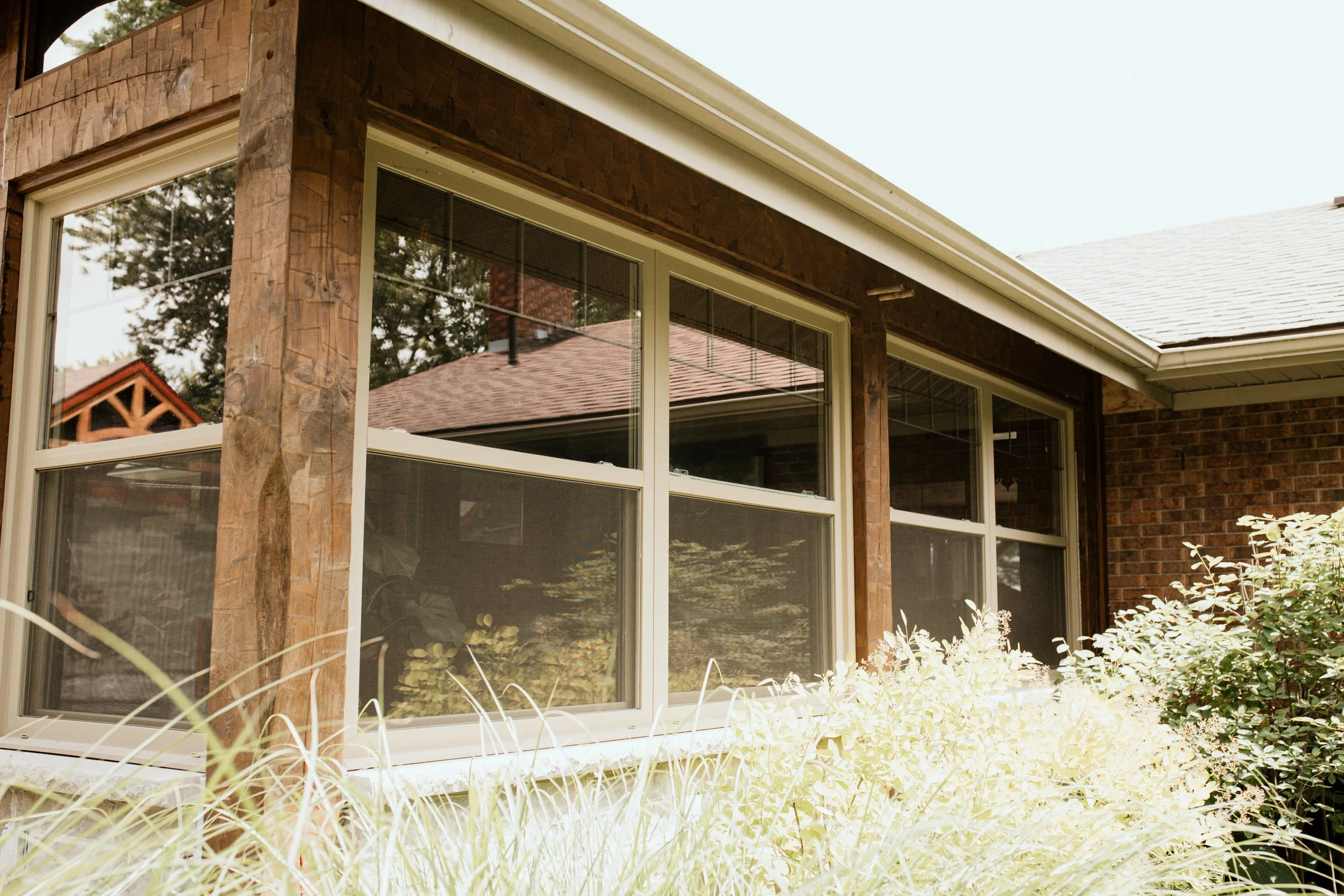 Close-up of a residential house's window and exterior wall with wooden trim and brick construction, surrounded by garden bushes and plants.