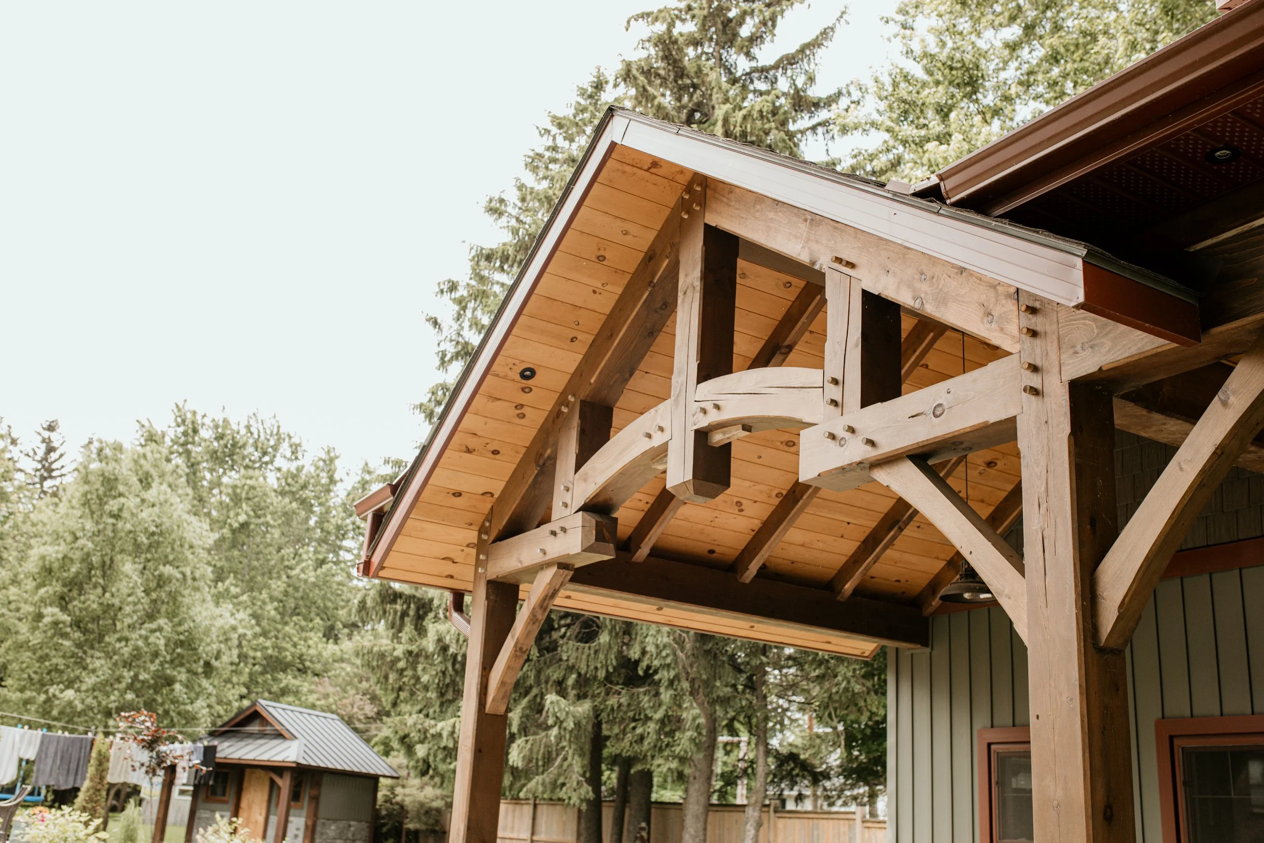 Part of a house's roof with exposed wooden beams and a new timber frame under construction, surrounded by trees.
