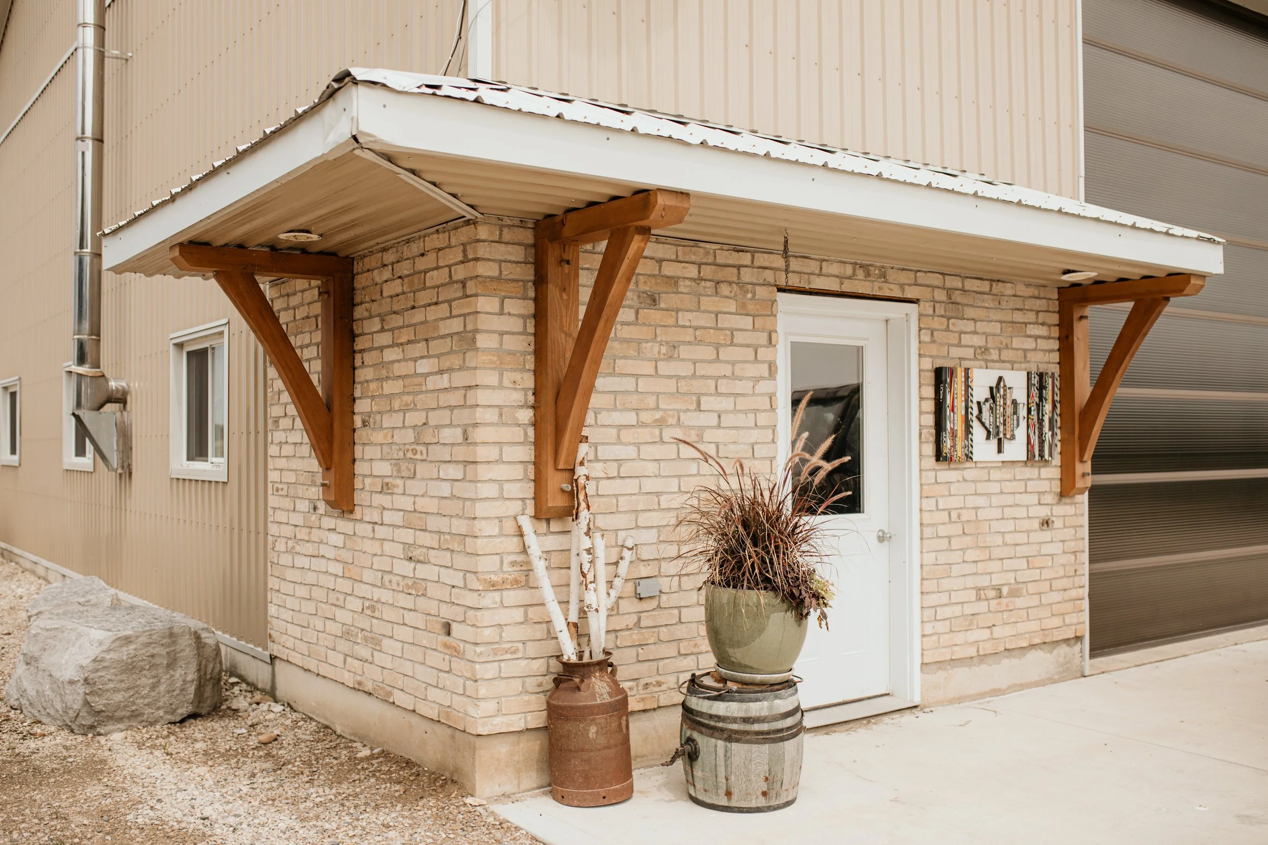 Small brick building with a white door, a decorative wall hanging, and two potted plants outside.