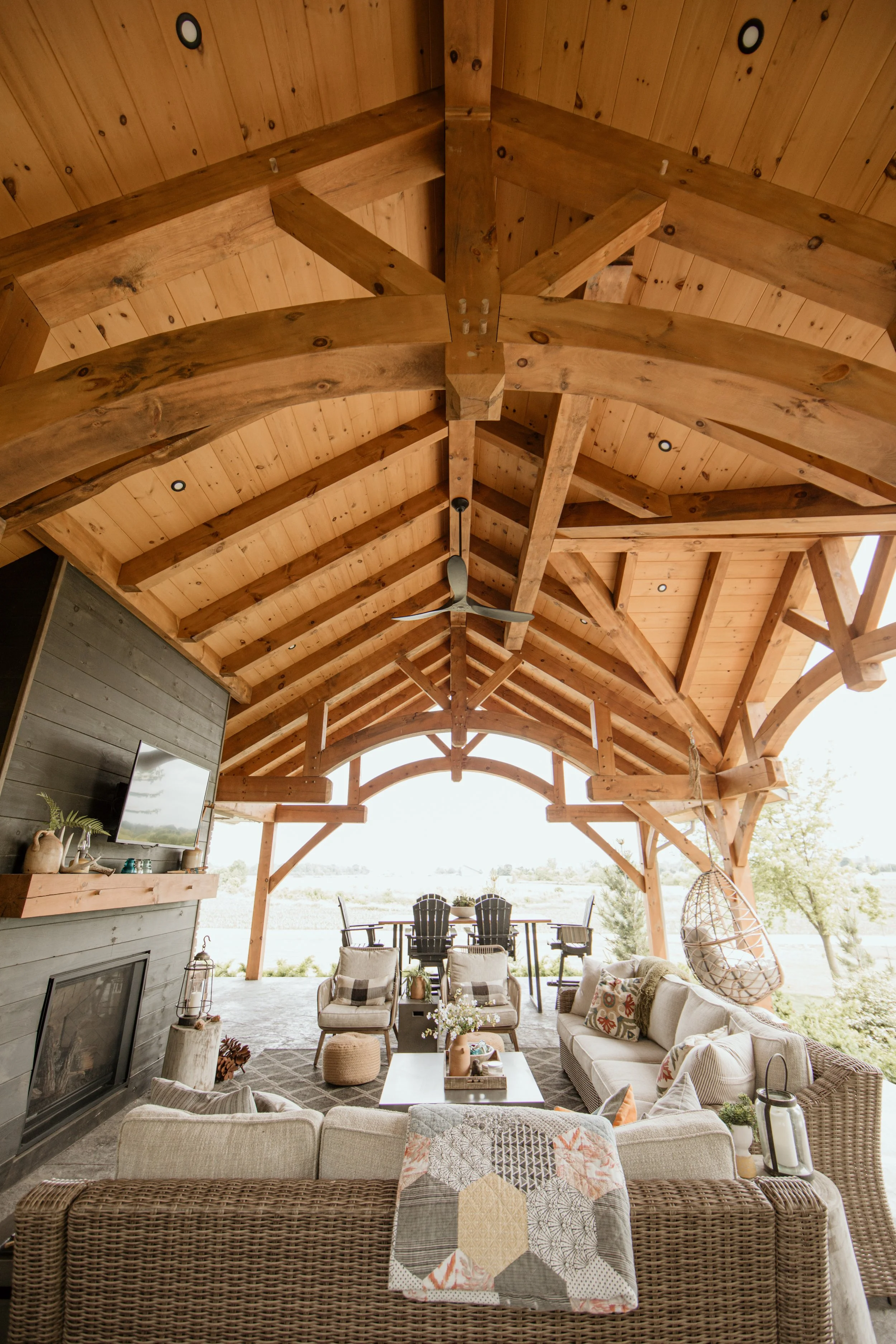 A spacious covered outdoor living area with wooden beams and ceiling, furnished with sofas, chairs, and a coffee table, overlooking a natural landscape.
