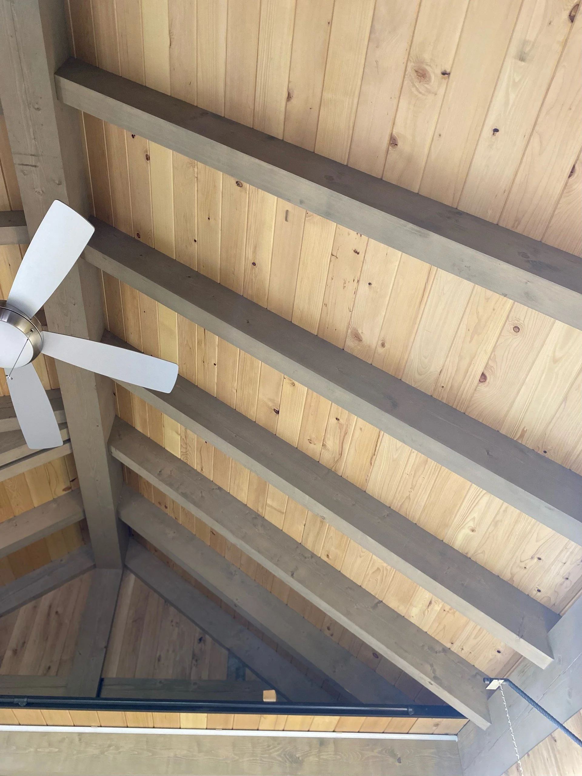 View of a ceiling with wooden beams, wooden paneling, and a ceiling fan with white blades.