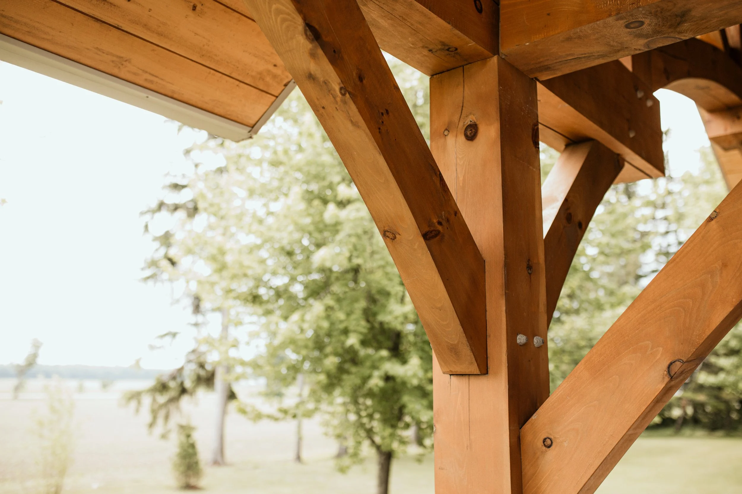 Close-up of wooden beams forming a corner of a wooden structure, with green trees and a field in the background.
