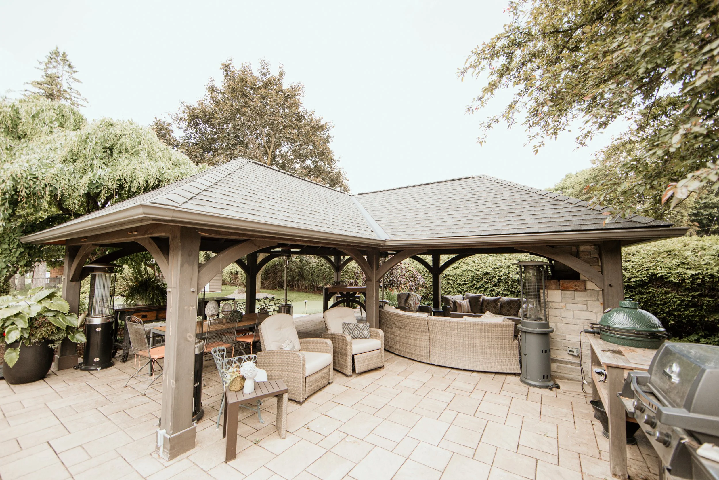 Outdoor patio with a wooden pergola, seating area with beige wicker sofa and armchairs, dining table with chairs, tall heaters, and a green barbecue grill, surrounded by trees and green hedges.